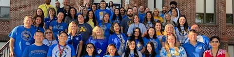 Group of adults and students in blue shirts, standing and sitting on steps outside a brick building.