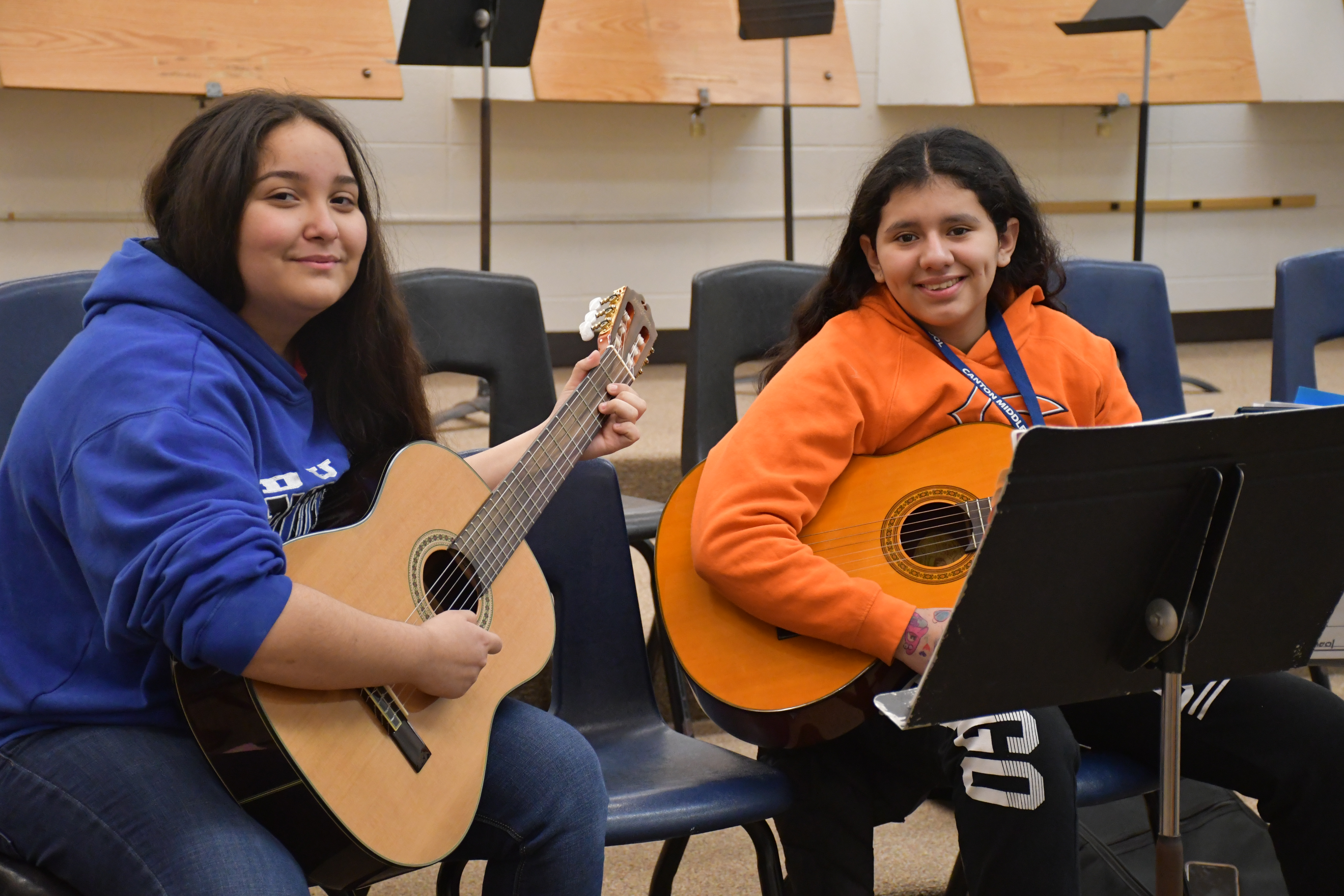 Two girls seated, one with a blue hoodie, the other in an orange shirt, playing guitars. Behind them, chairs and music stands.