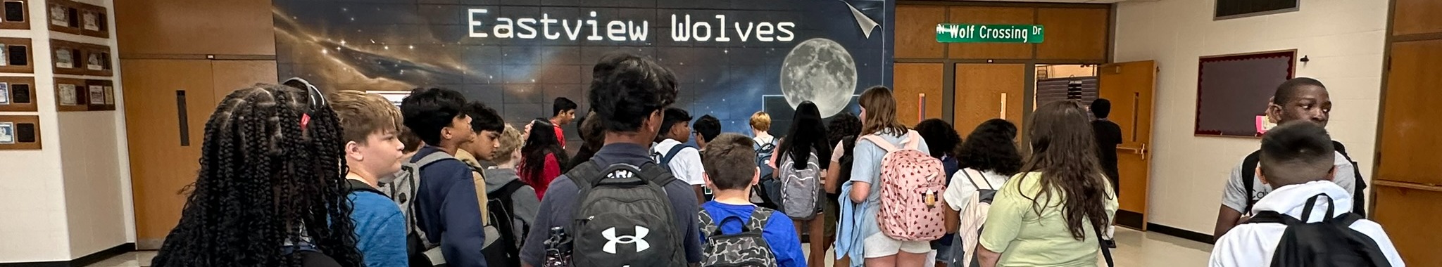 A hallway with students standing, some wearing backpacks. One person holds a sign that reads Eastview Wolves.