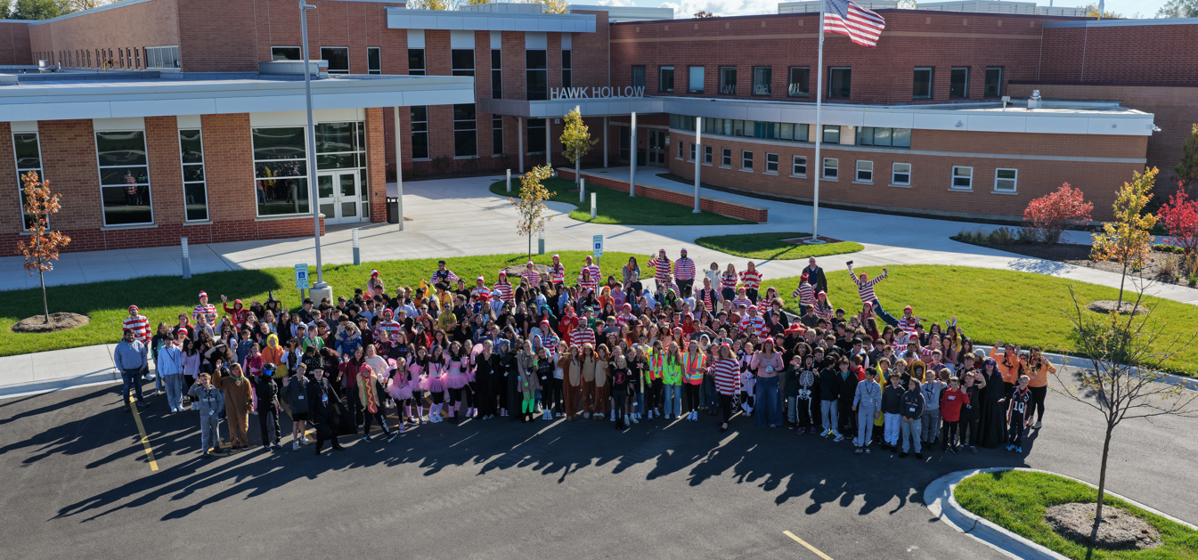 Many people stand in front of a school building, with trees, plants, and an American flag visible.