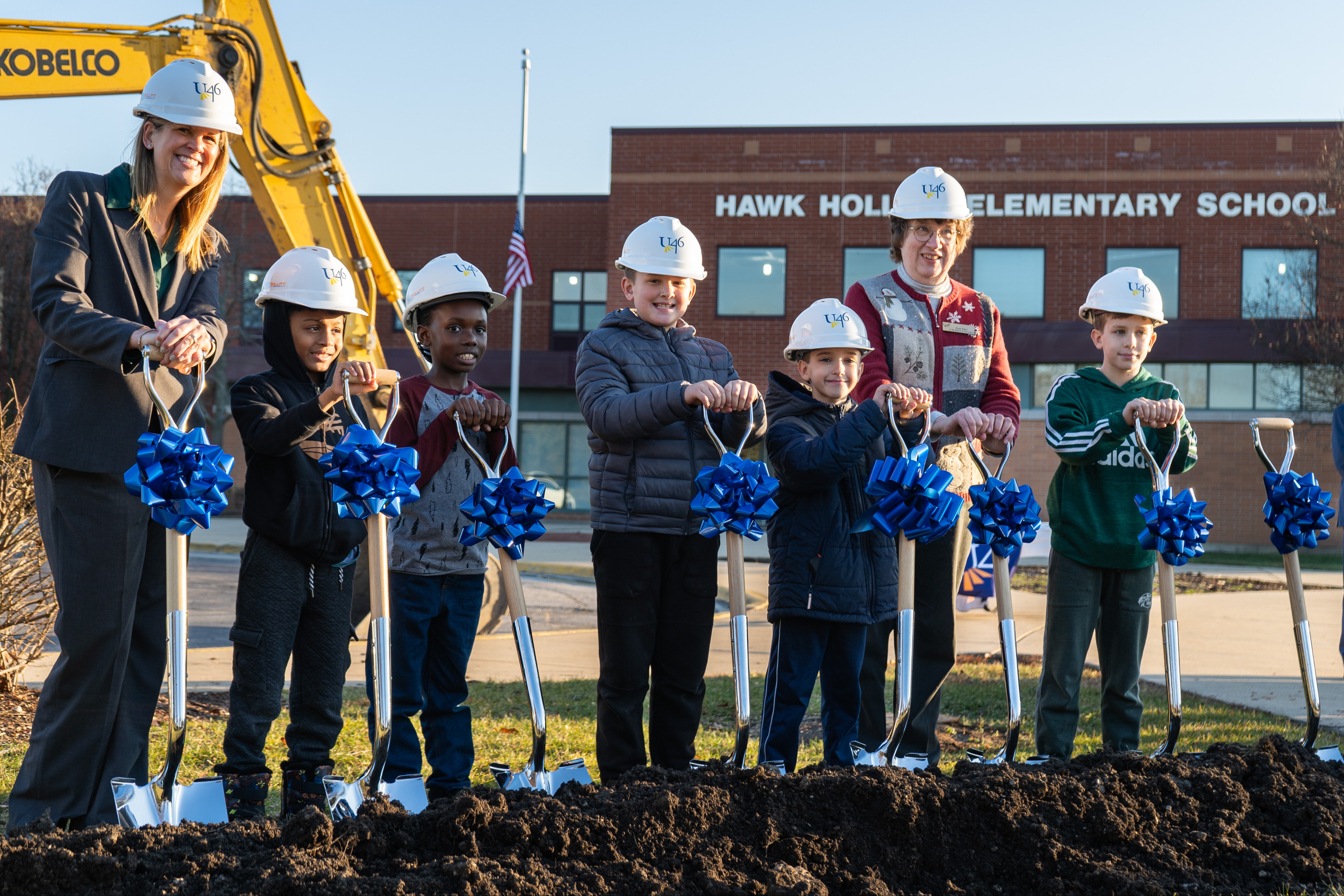 People, adults and children, stand around a construction site with shovels. Helmets and shovels are decorated with blue ribbons.