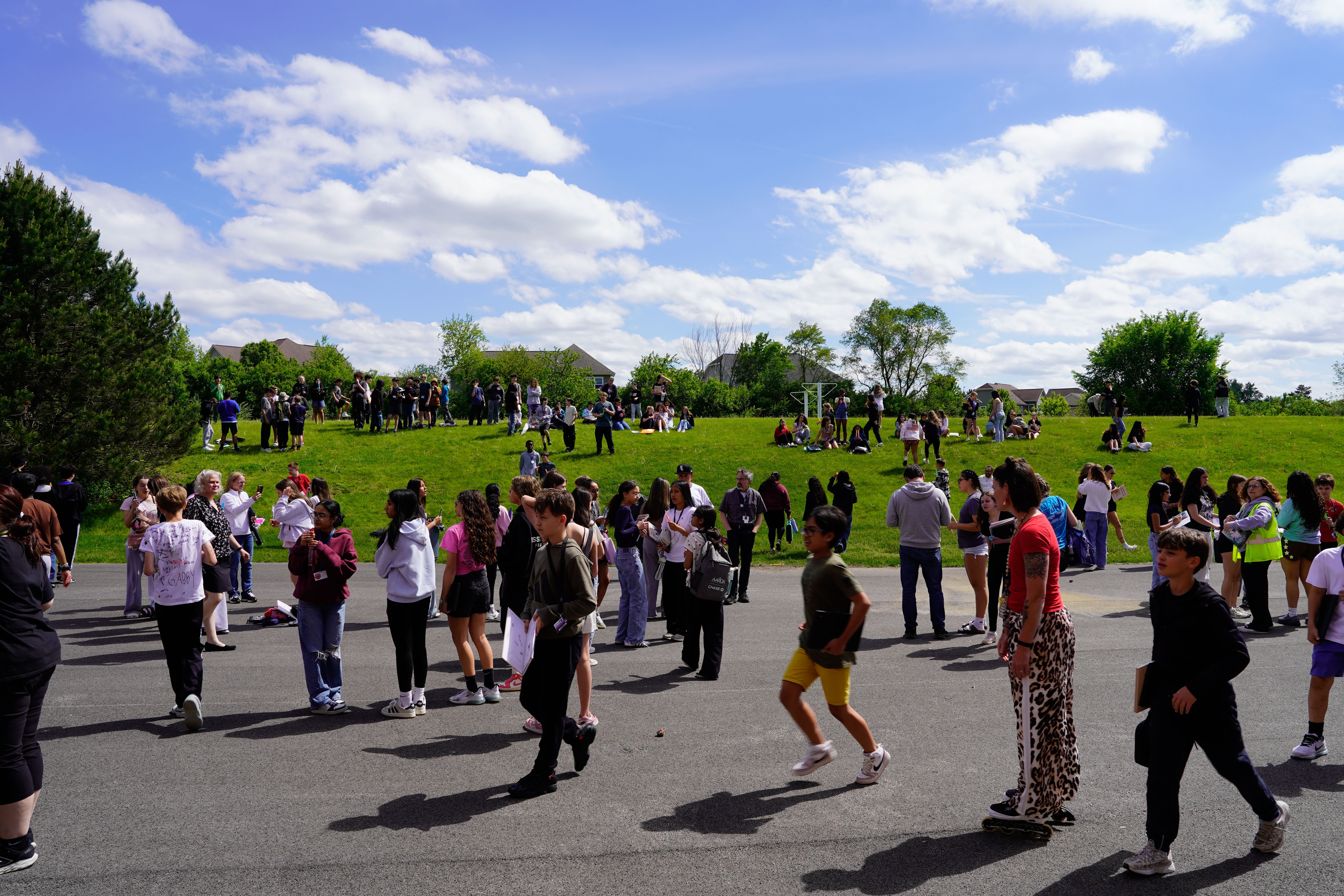 An open area with people walking. Trees and grass surround the area. A clear blue sky with some clouds is in the background.
