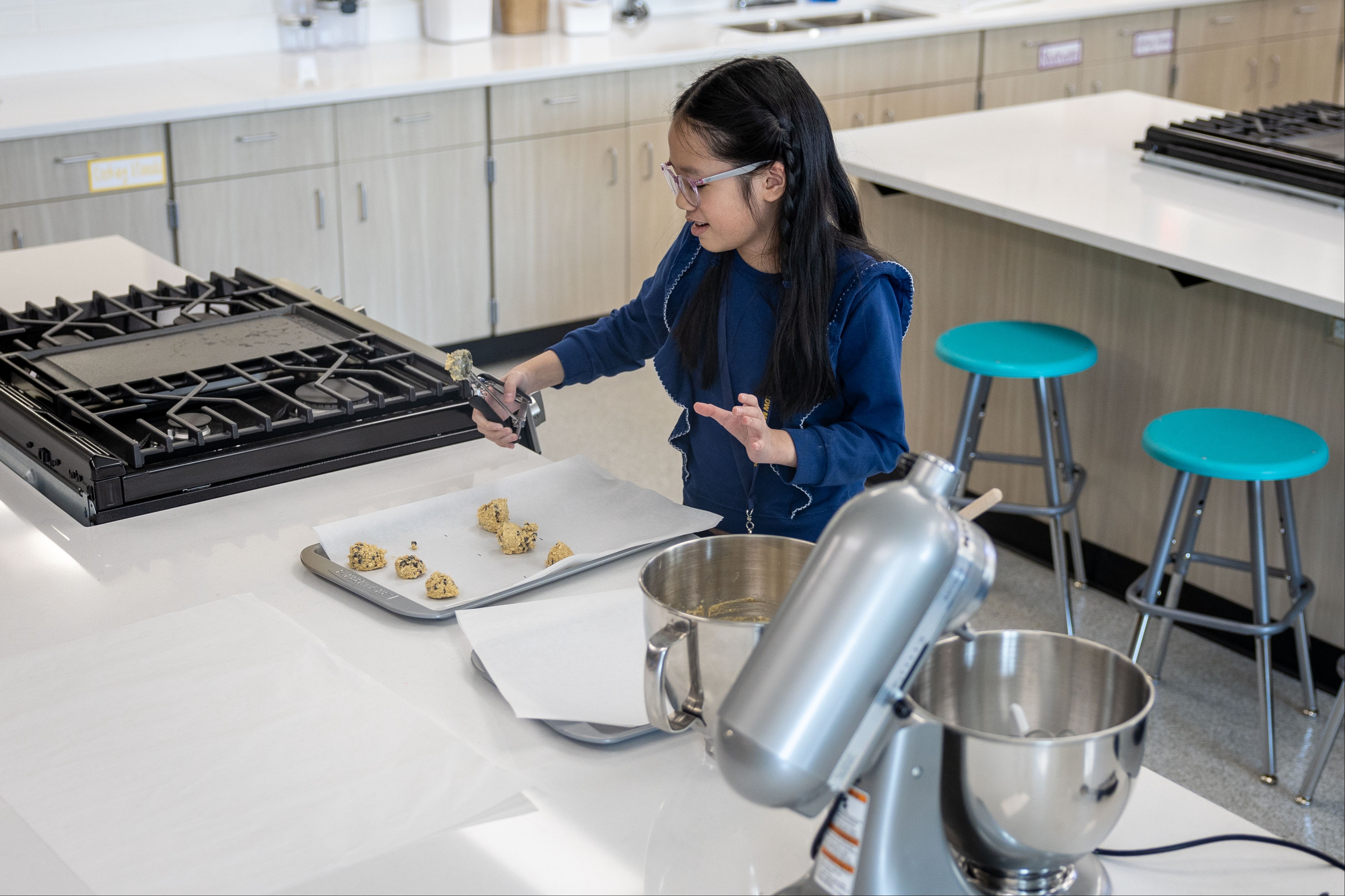 Child in a kitchen with glasses holding a cookie cutter and cookies on a baking sheet.