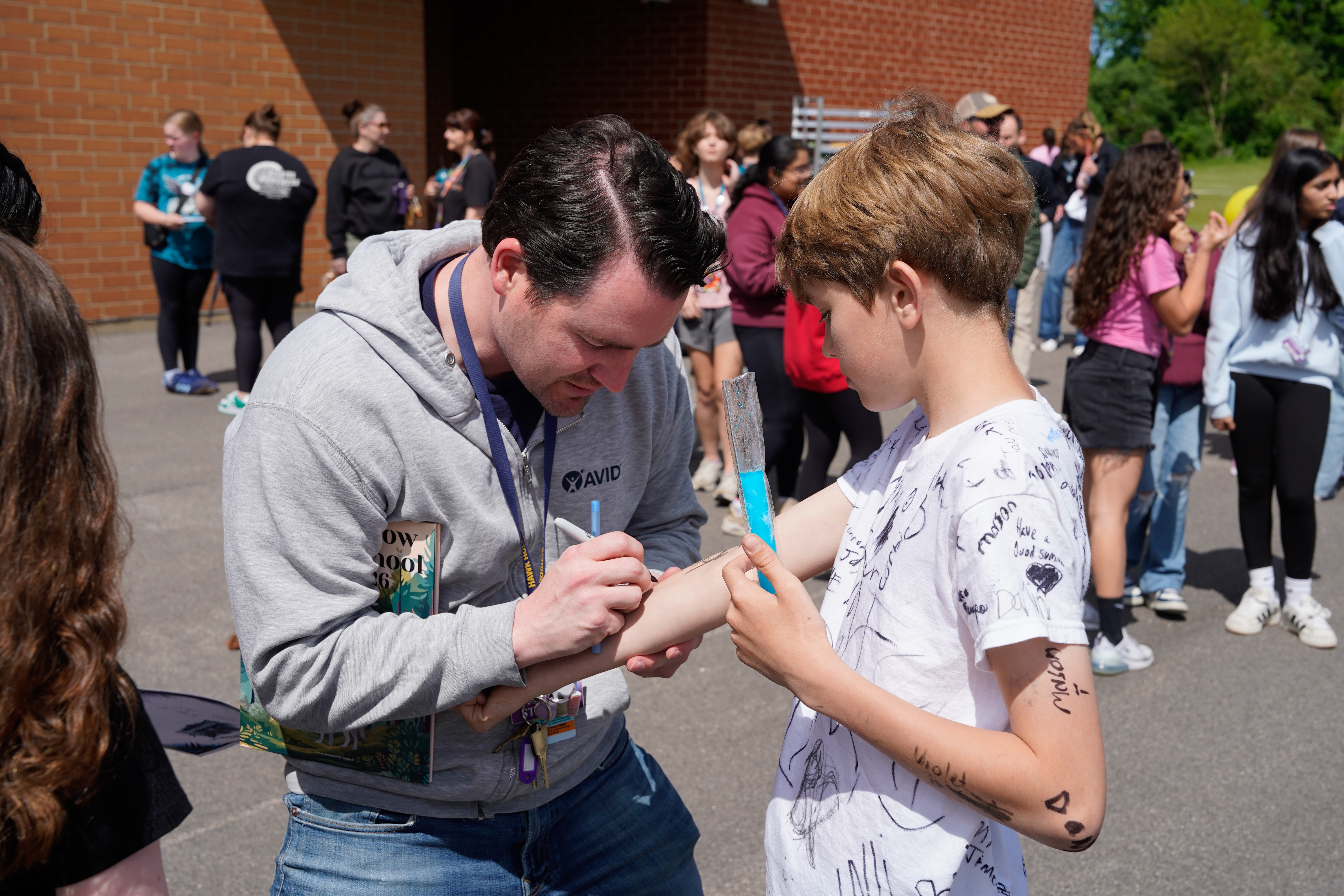 A man helps a young boy write on his arm in a school parking lot with many people.