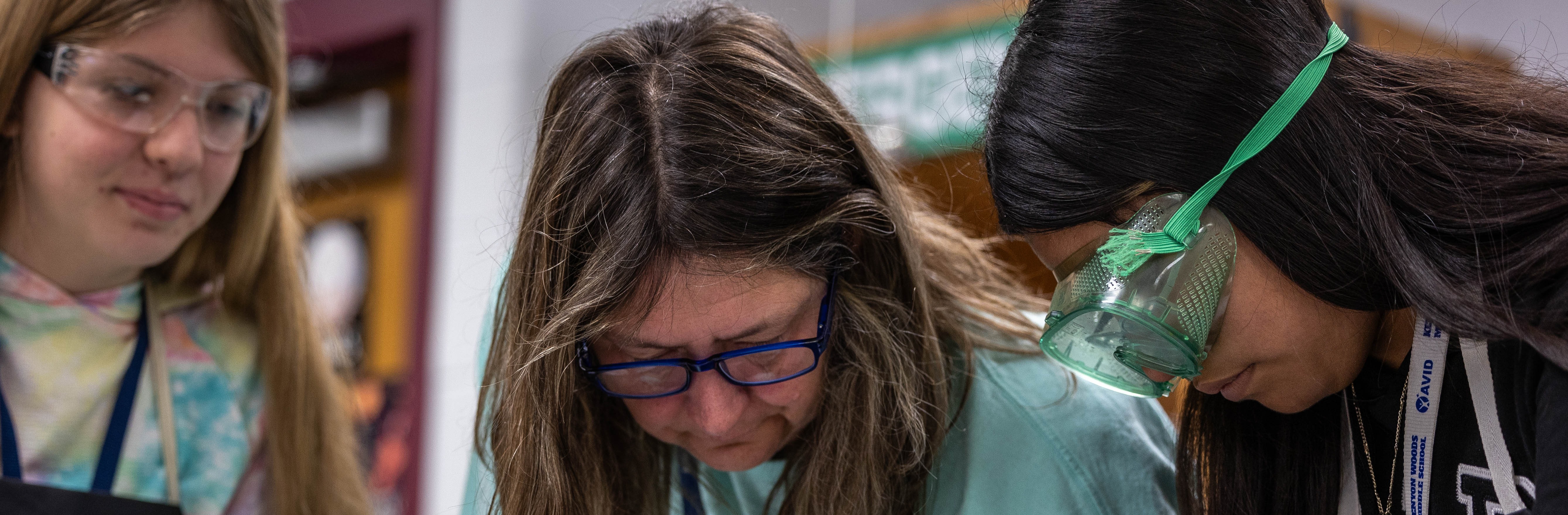 Three individuals with glasses examine a clear glass cylinder in a room. The person on the left wears a tie-dye hoodie.