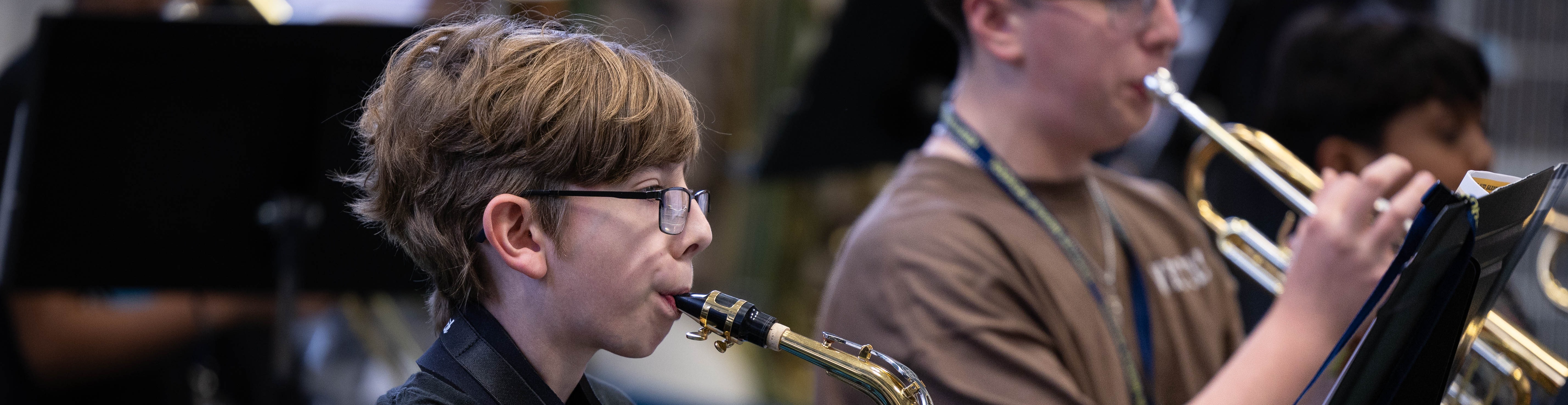 Two boys, one in black and the other in brown, play saxophone and trumpet in a band setting.