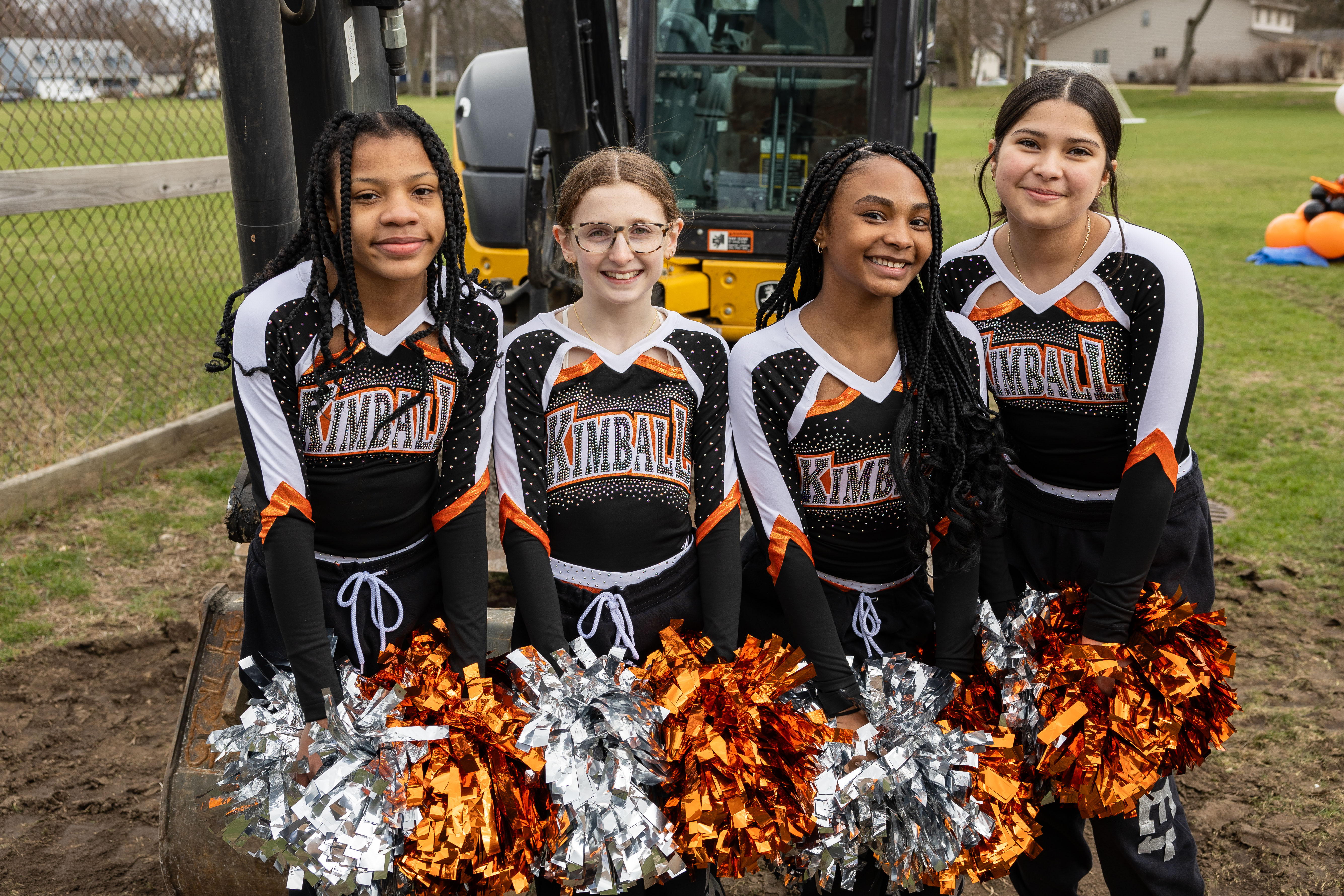 Four cheerleaders, wearing black, white, and orange uniforms, stand next to a yellow construction vehicle. They hold silver and orange pom-poms.