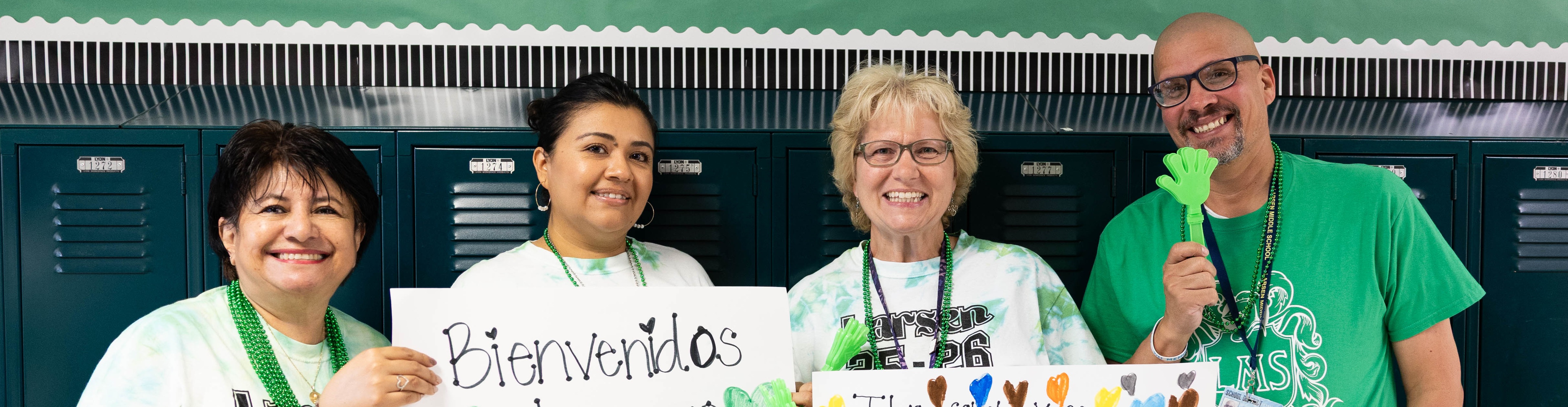 Four individuals stand behind lockers, smiling. They hold a sign with rainbow hearts and another with colorful artwork.