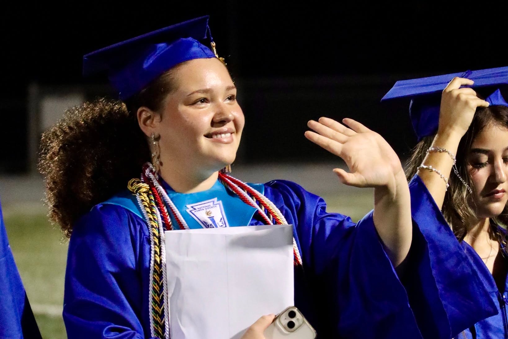 Female graduate in blue graduation robe waving and smiling.