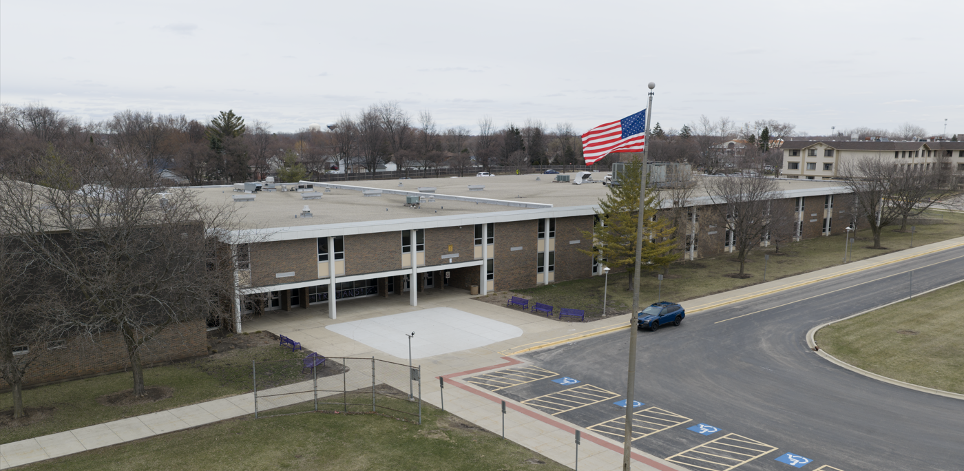 A school building with a flagpole in front. A car is parked on the side of the road.