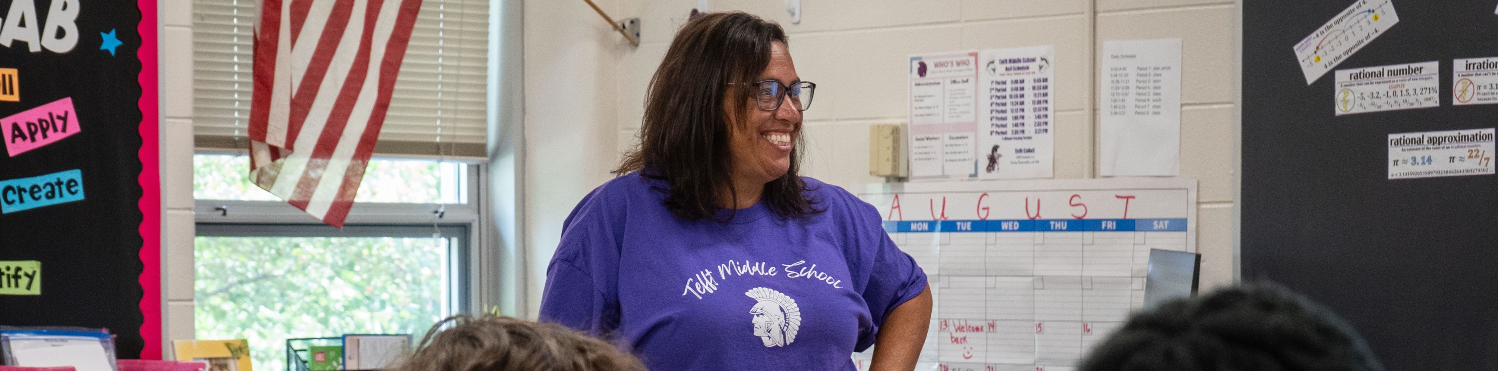 A teacher in a purple shirt stands near a classroom window with an American flag. Students sit at tables.