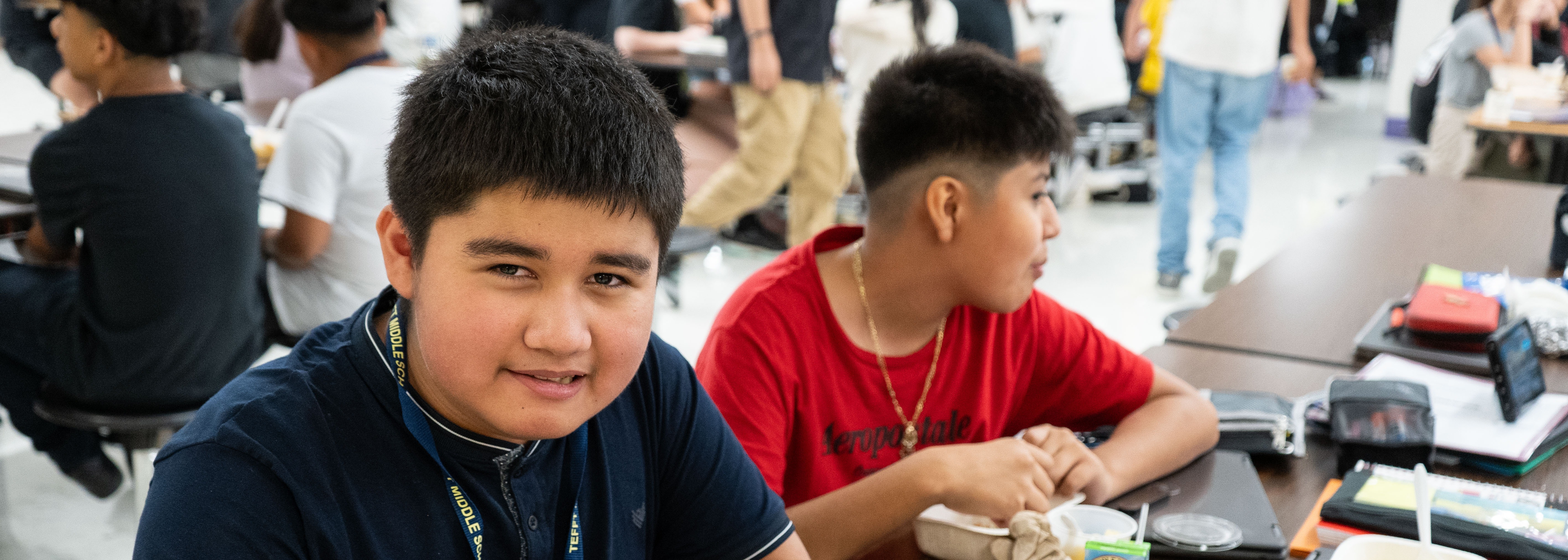 Two students seated at a table, one eating a sandwich, with various food items and school supplies around them.