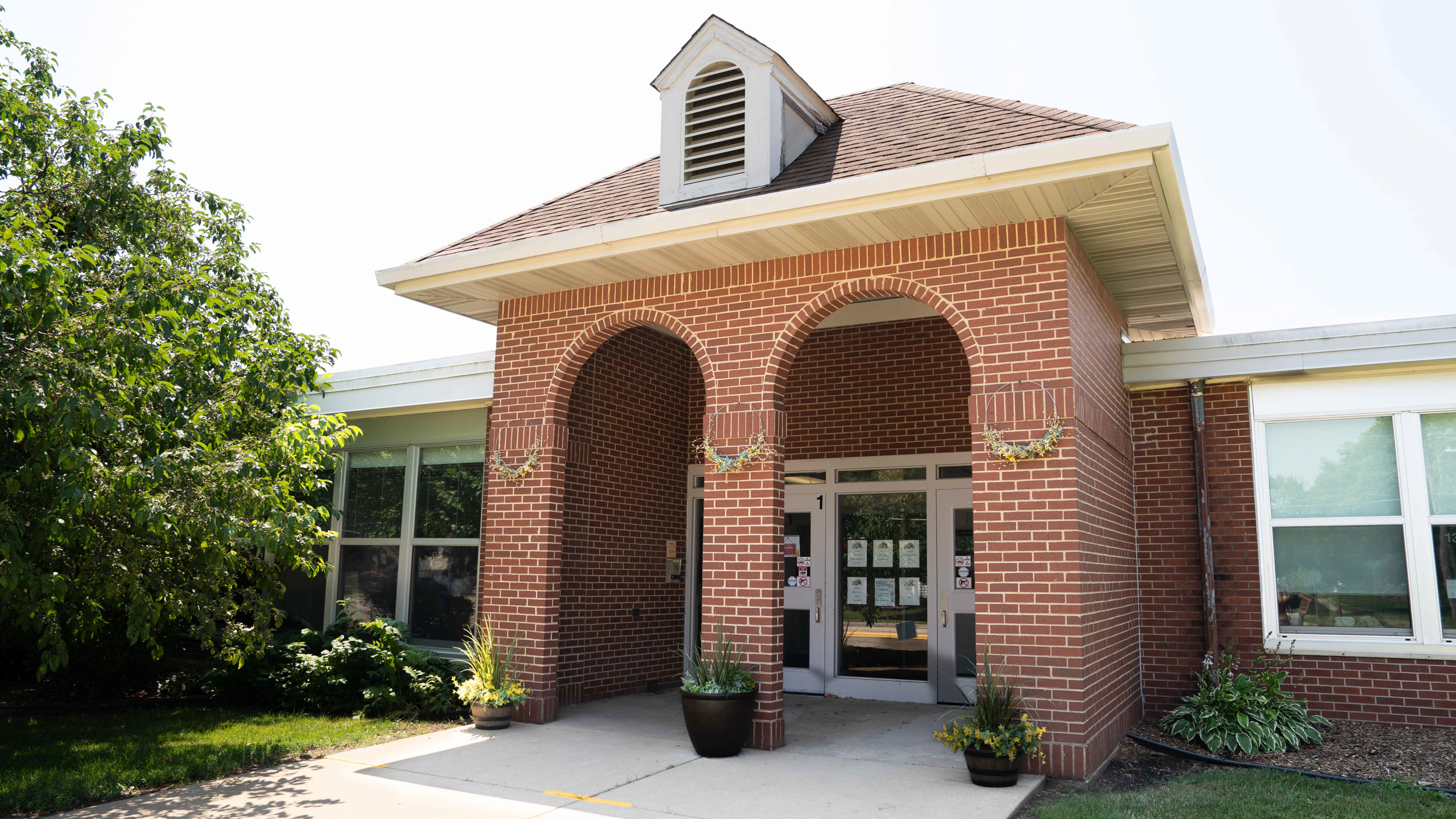 Brick building entrance with arched doorways and large windows. Potted plants sit at the front.
