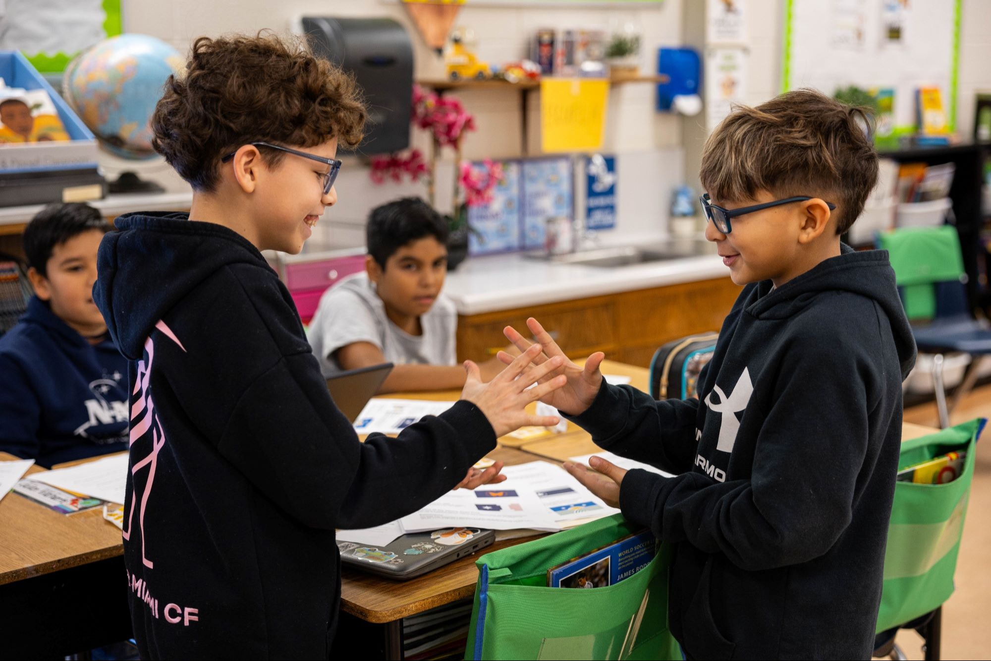 Two children shake hands at a table in a classroom with other students seated behind them.