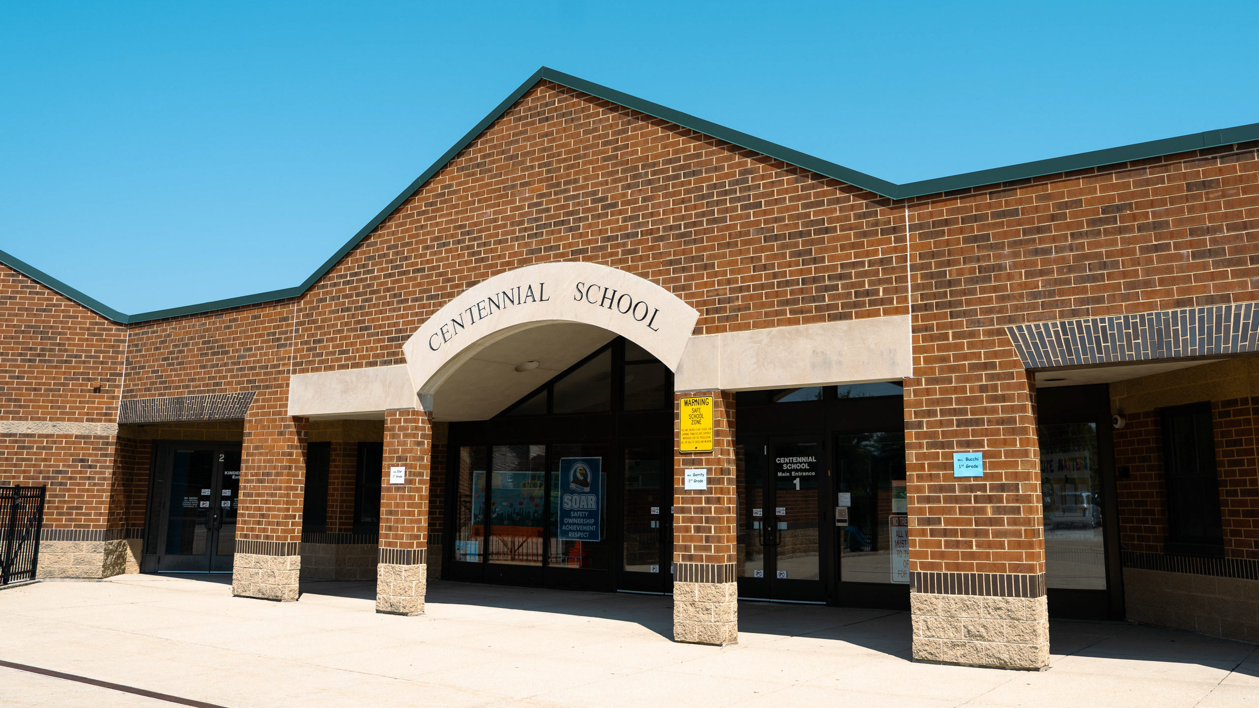Entrance of a brick school building with arched doorway, "Centennial School" sign, and glass doors.