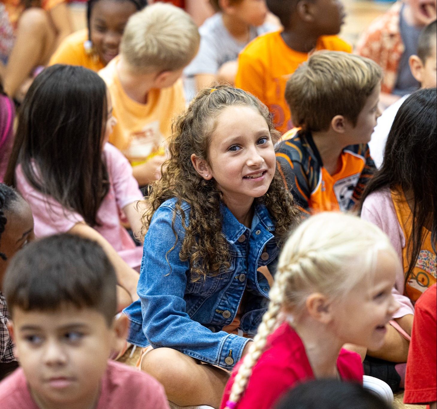 Several children seated, some smiling and others attentive, in a classroom with a blurred background.