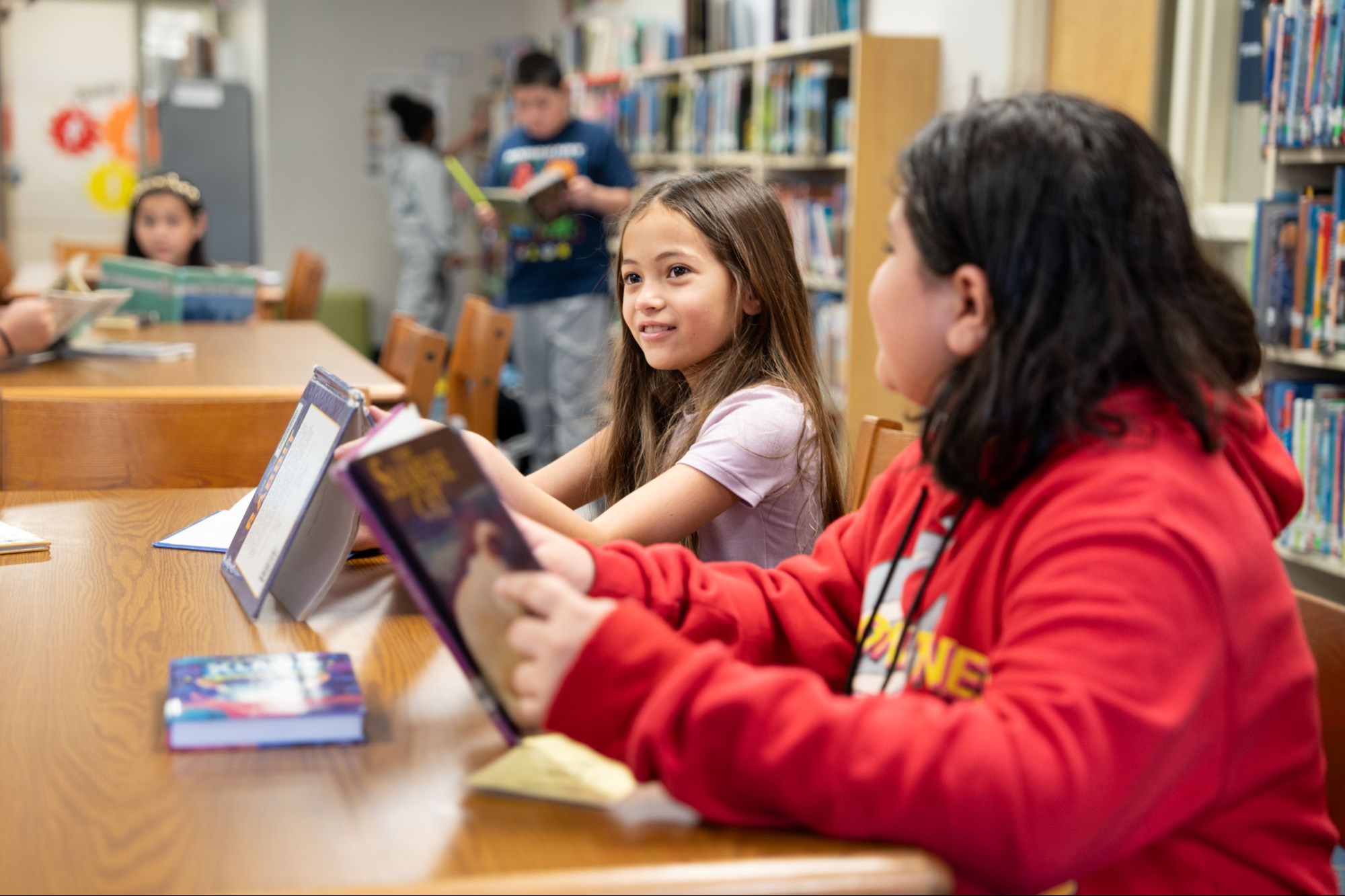 Two children sit at a table in a library. One holds a book, the other reads aloud. Shelves full of books are in the background.