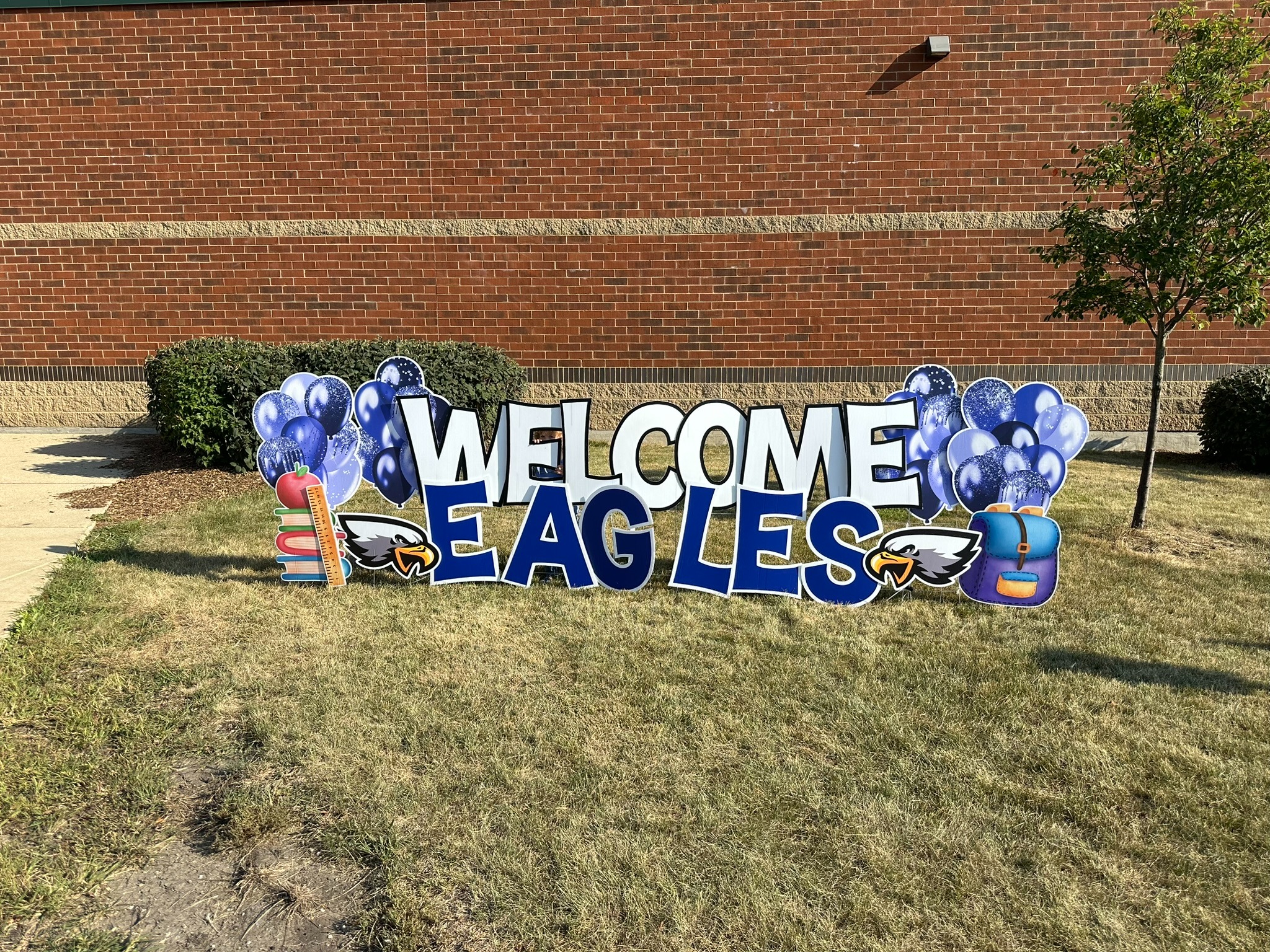 Grassy area with a sign saying "Welcome Eagles" in blue and white letters. A brick building is in the background.