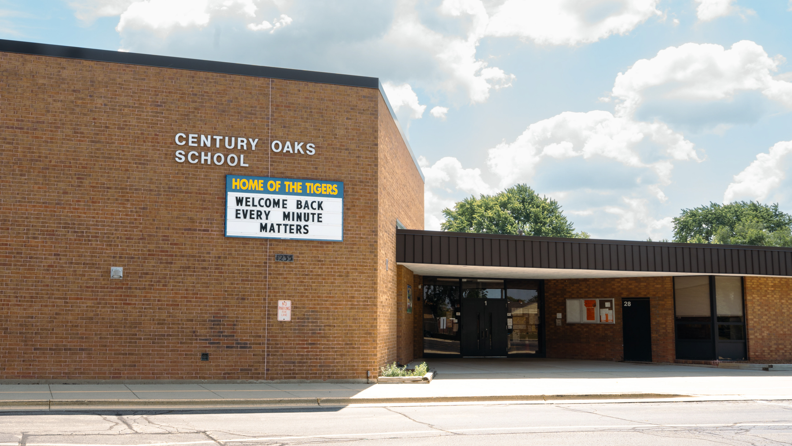 A building with a sign reading "Century Oaks School" and "Welcome Back Every Minute Matters" under a partly cloudy sky.