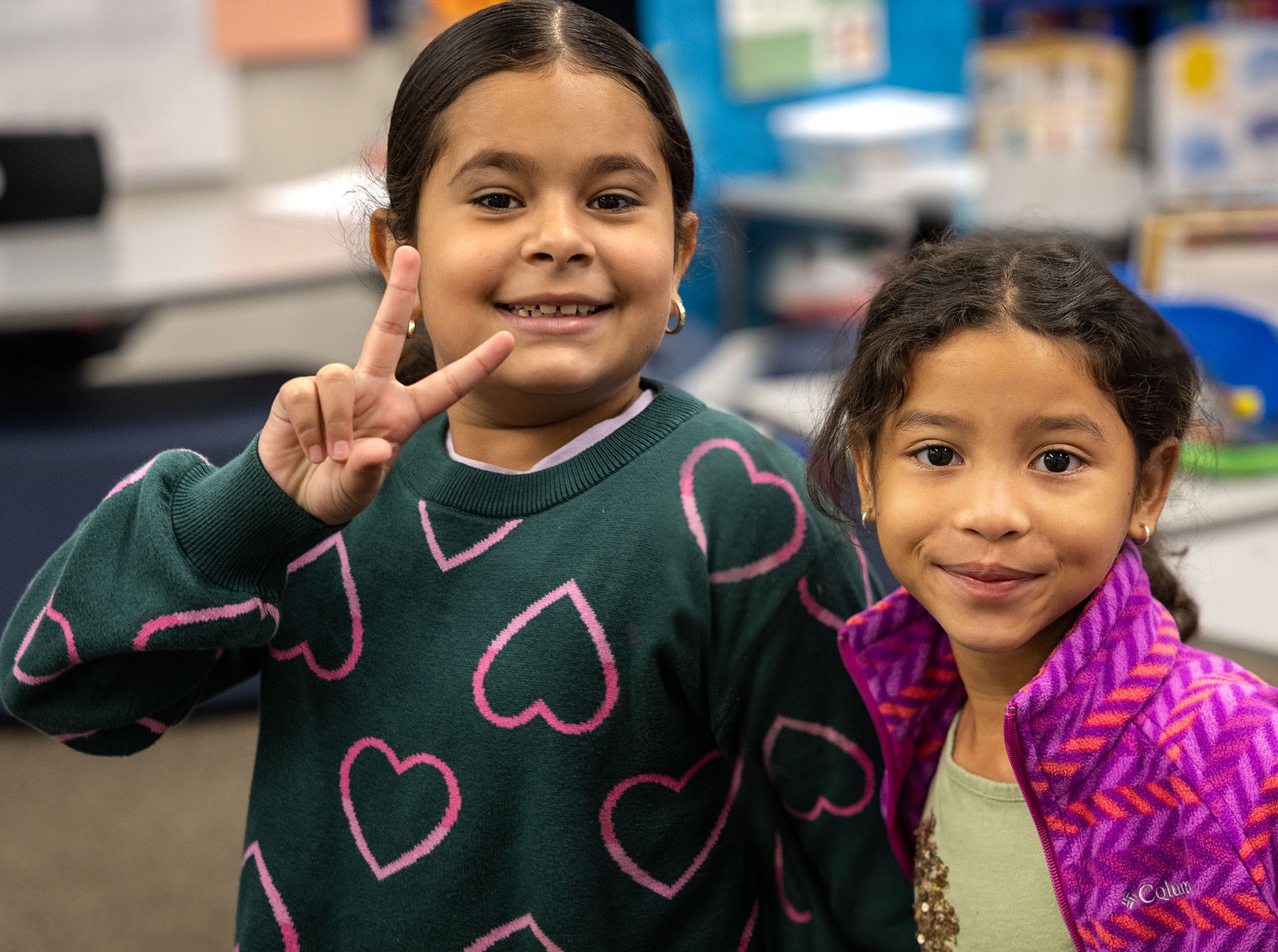 Two girls standing in a classroom. One girl makes a peace sign. Both have happy expressions.