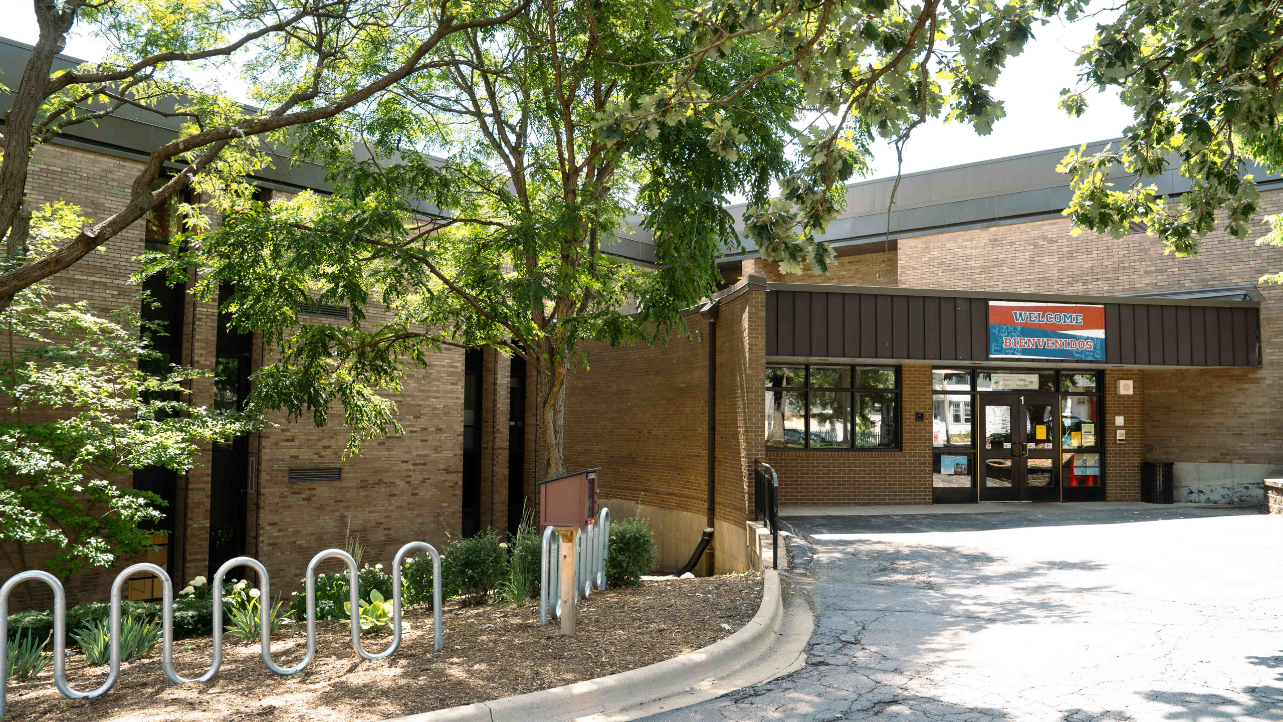 A building with brick walls and brown roofing. Trees and plants are in front of the building.