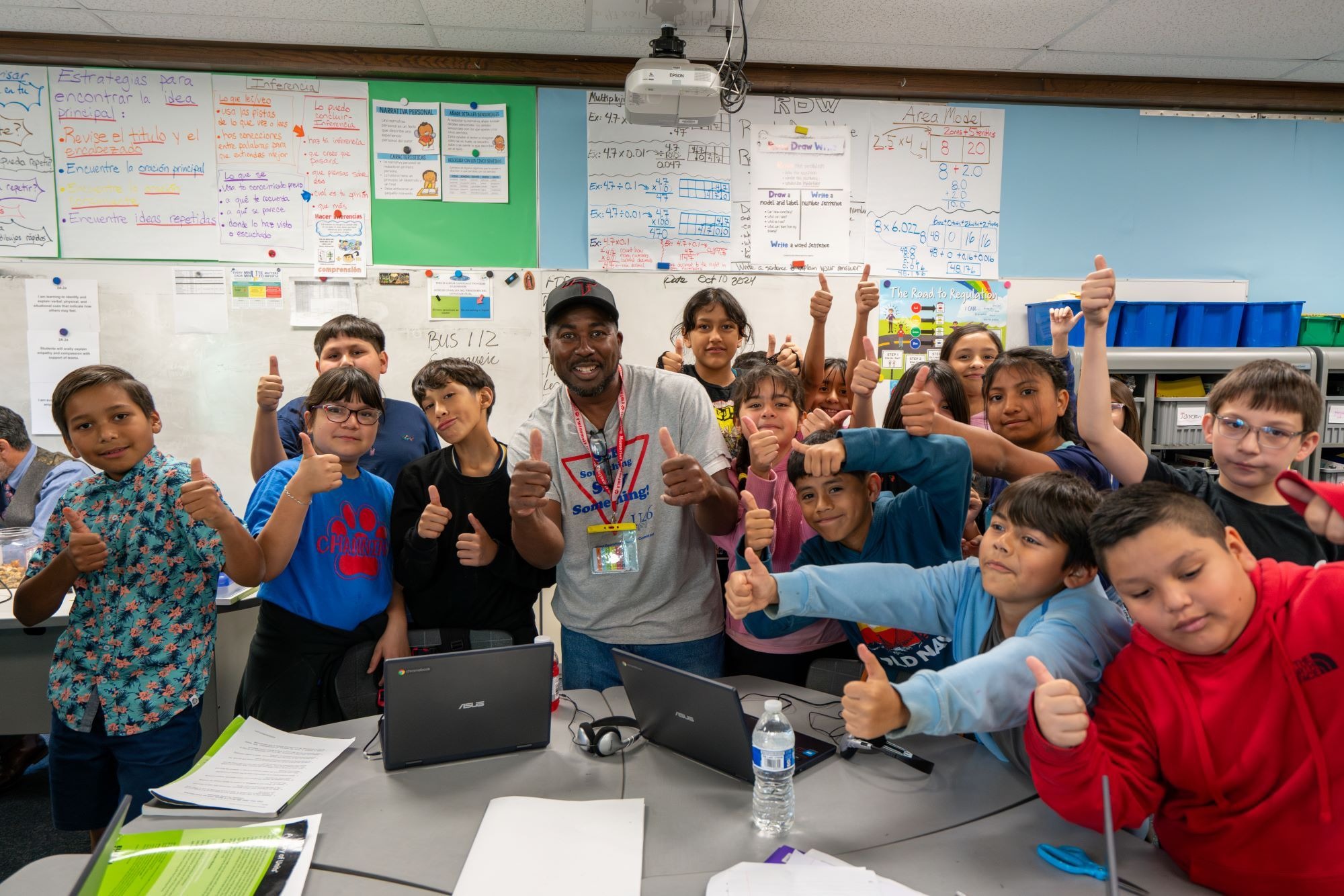 Kids and a man in a classroom, posing for a photo with thumbs up; laptops, papers, and a water bottle on the table.