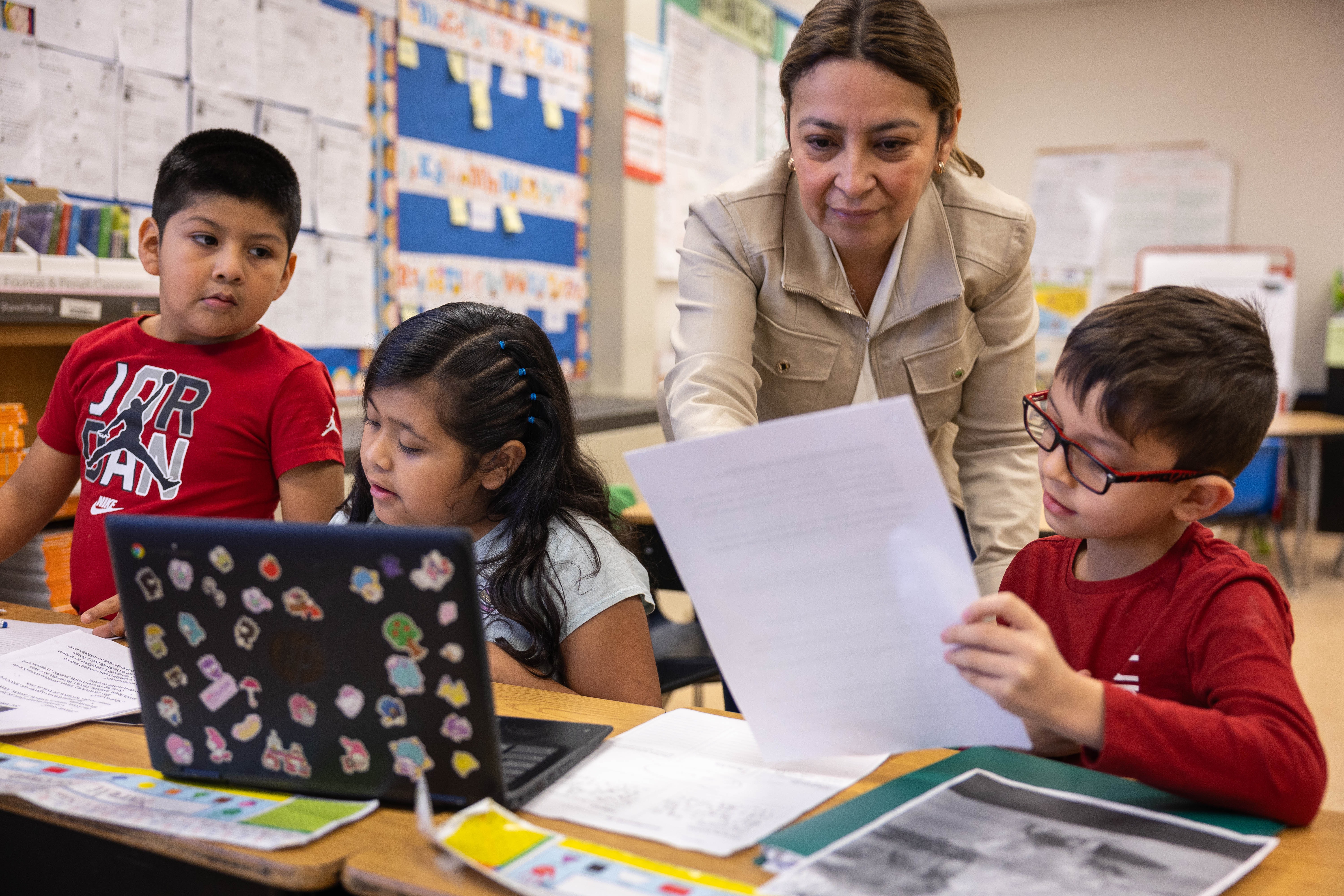 A woman in a beige blazer assists a boy in a red shirt with papers. A girl sits next to a laptop.