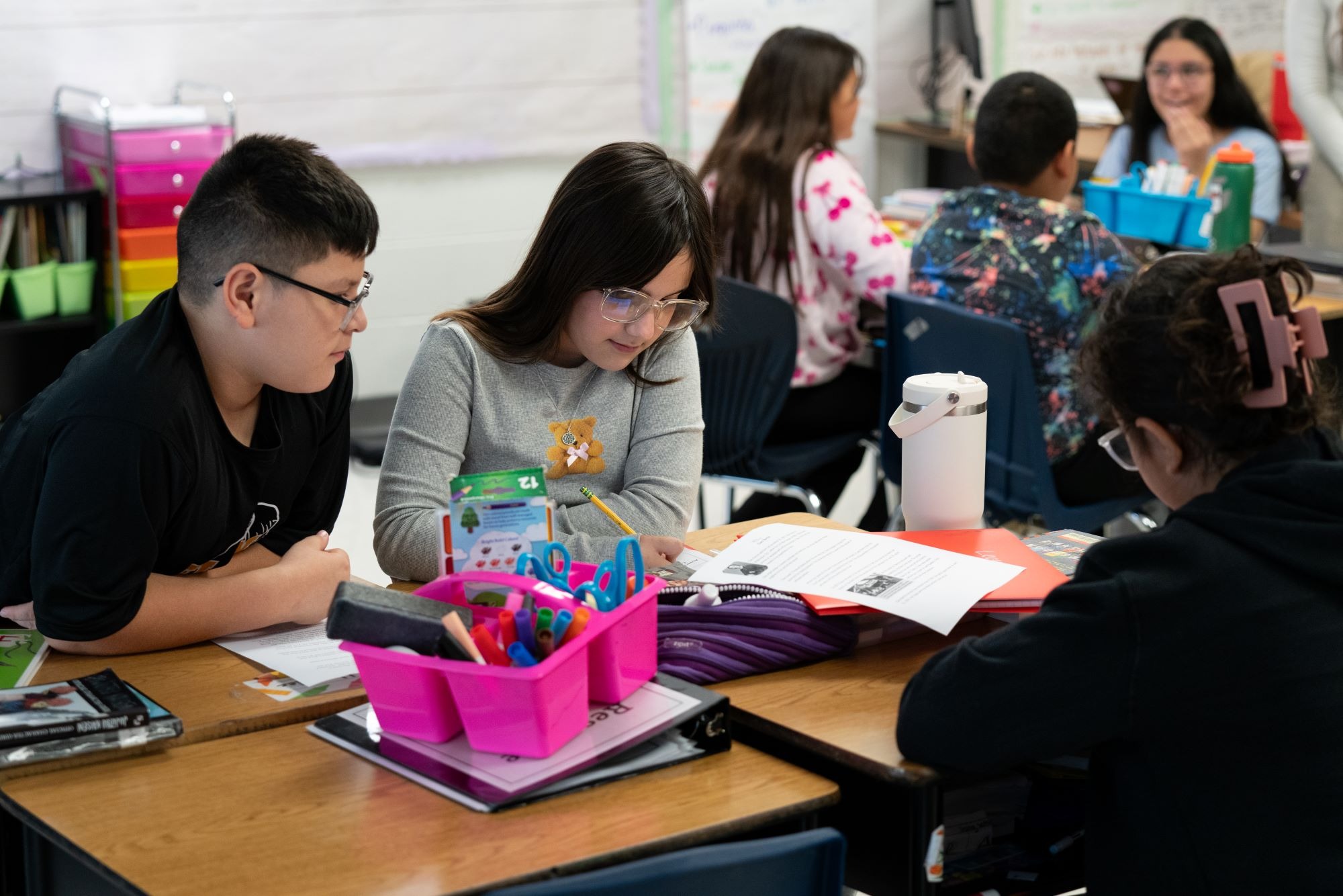 Students work in a classroom, seated at desks. Two students are looking at a worksheet.
