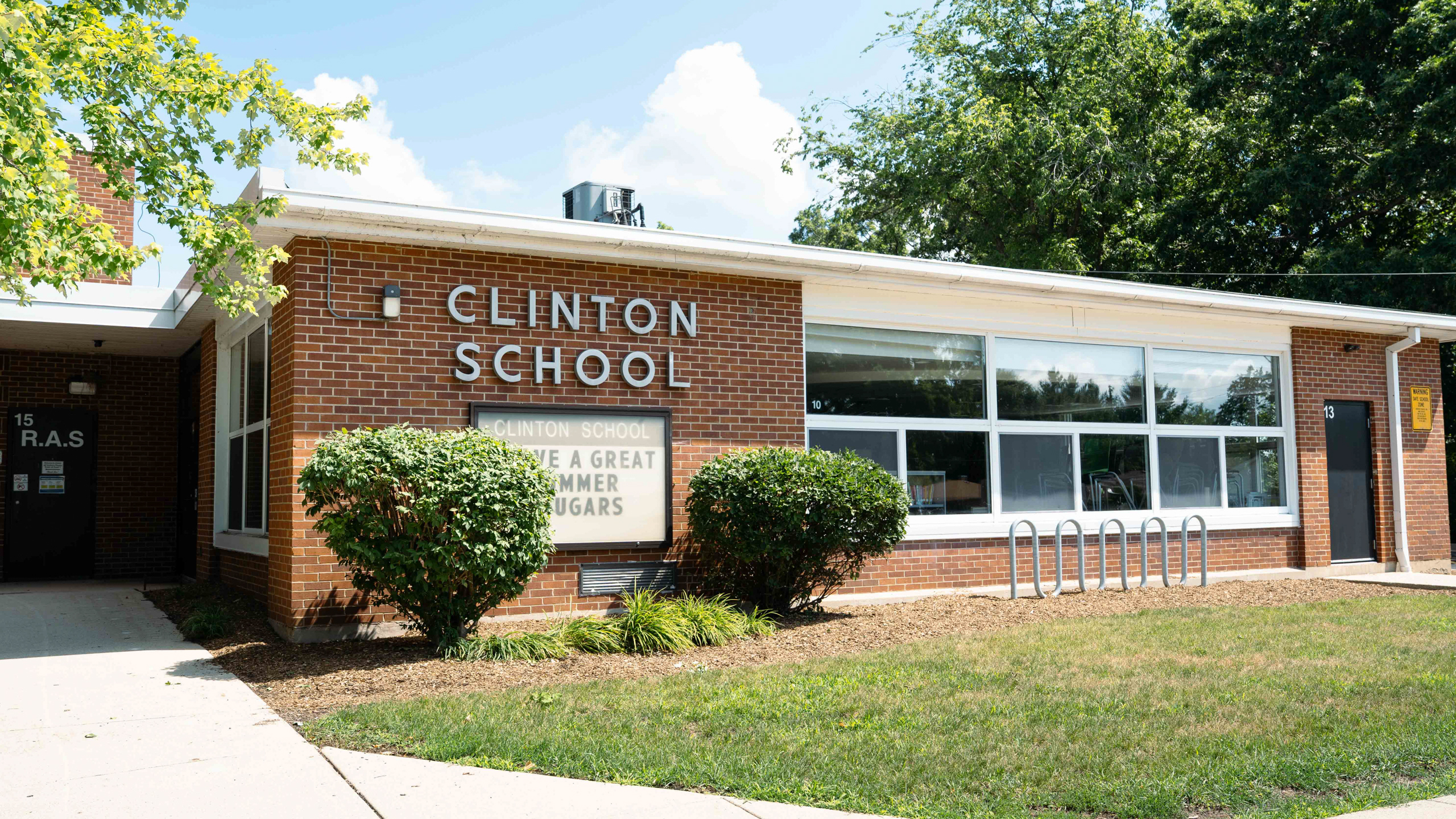 A building with "CLINTON SCHOOL" on the front. It has glass windows, a brick exterior, and bushes.
