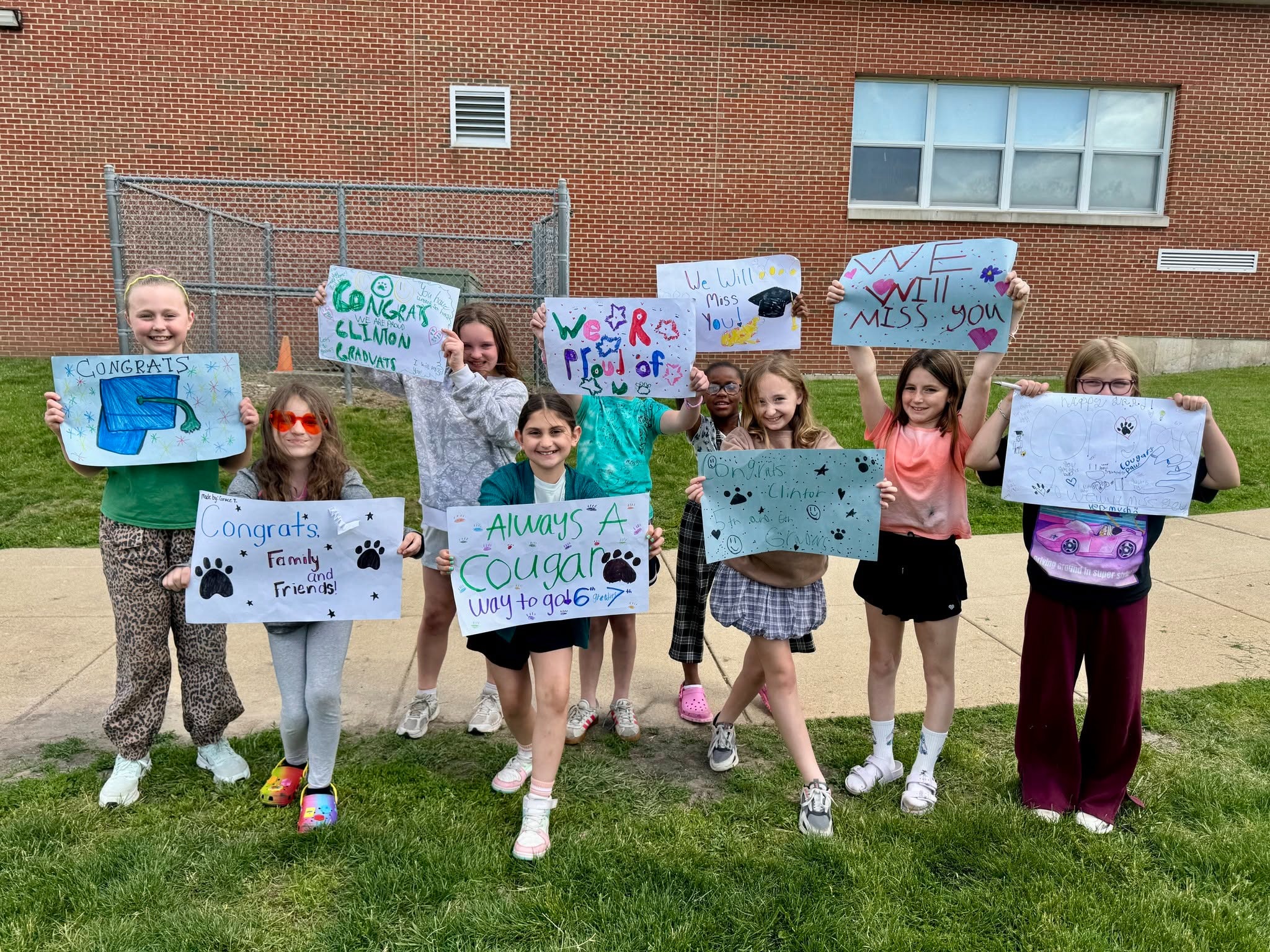 Group of children standing on grass holding signs with various messages in front of a brick building.