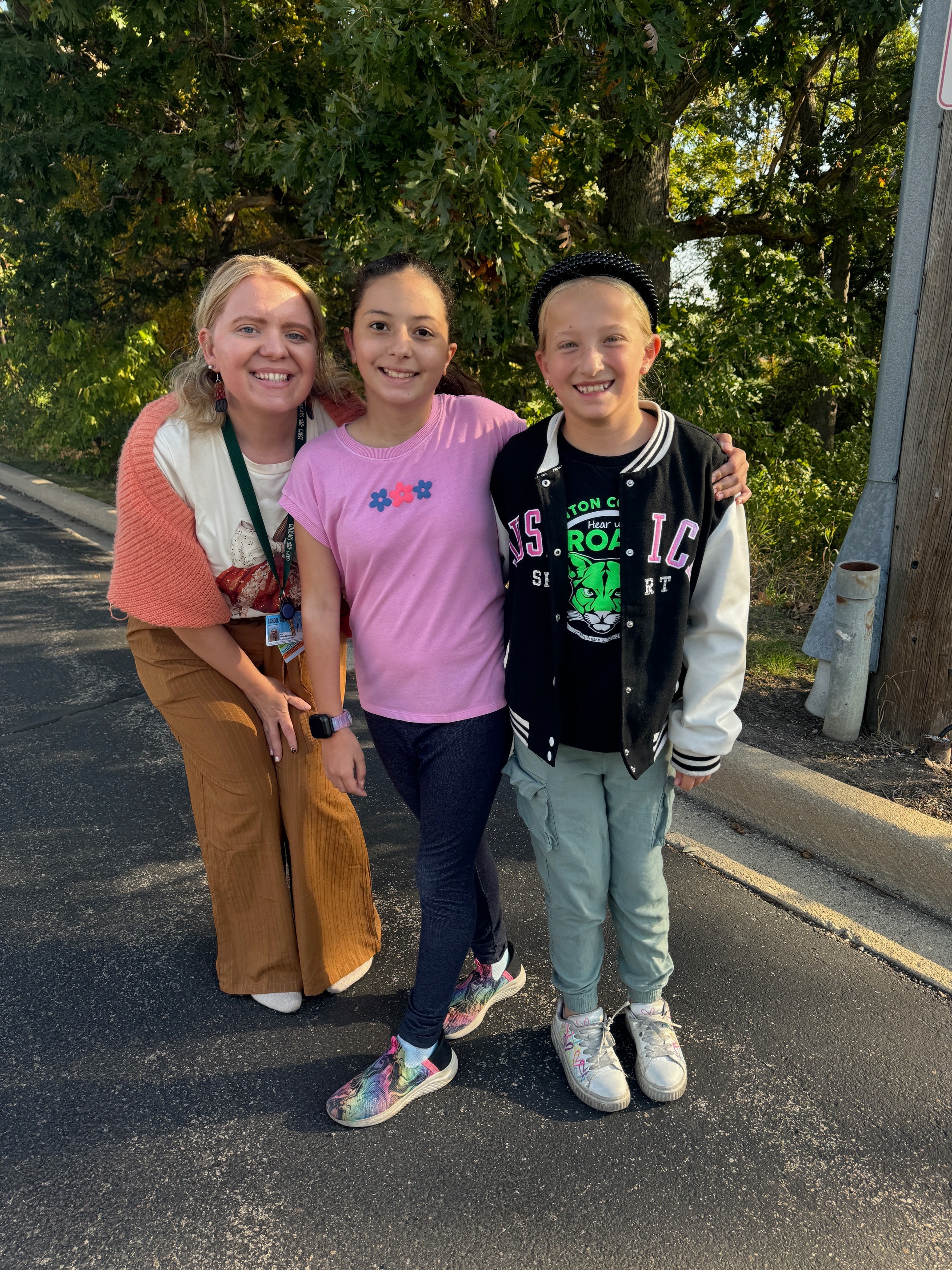 Three people pose on a street with trees and a utility pole in the background.