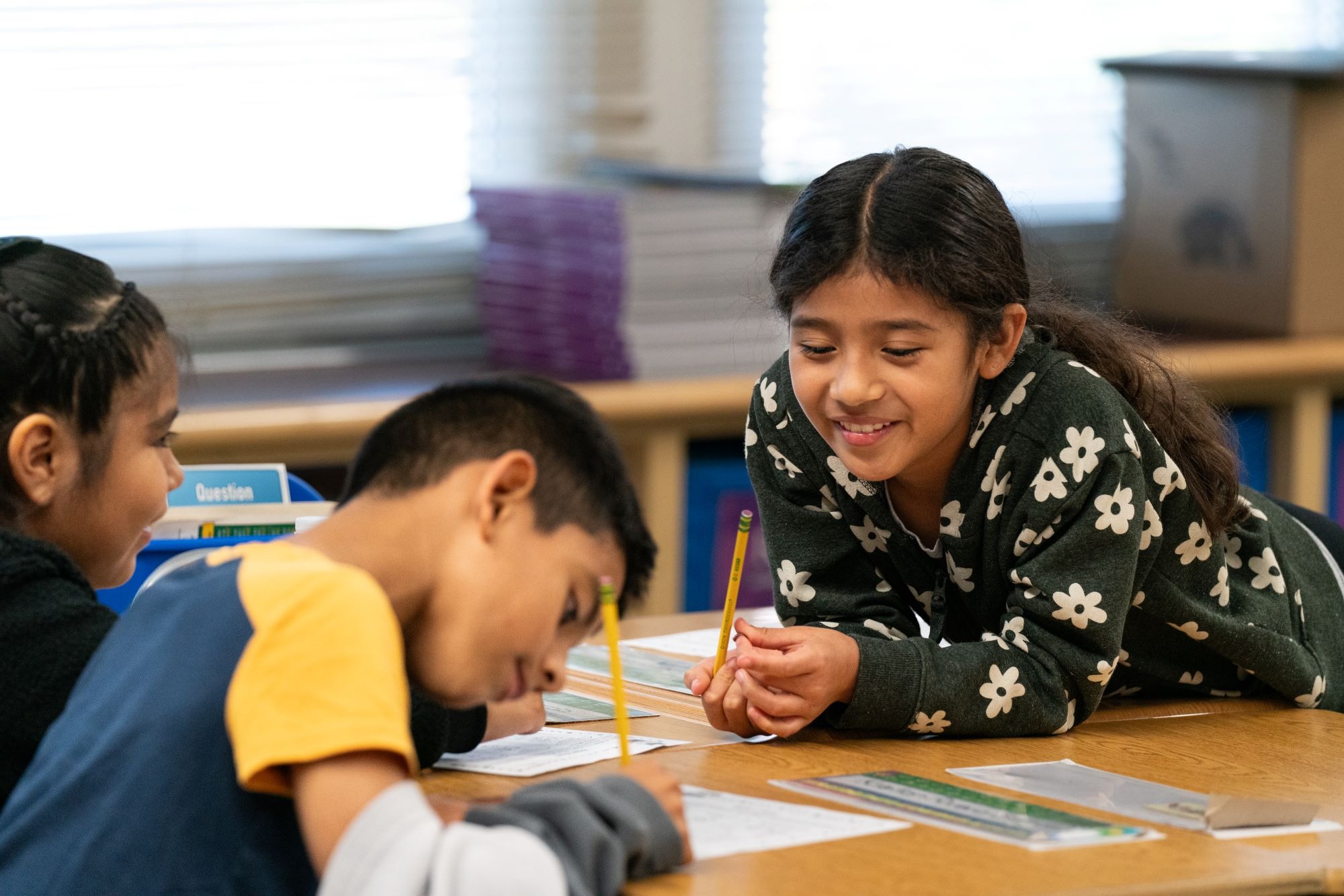 Children work on a project at a desk; one girl smiles at a boy who concentrates.