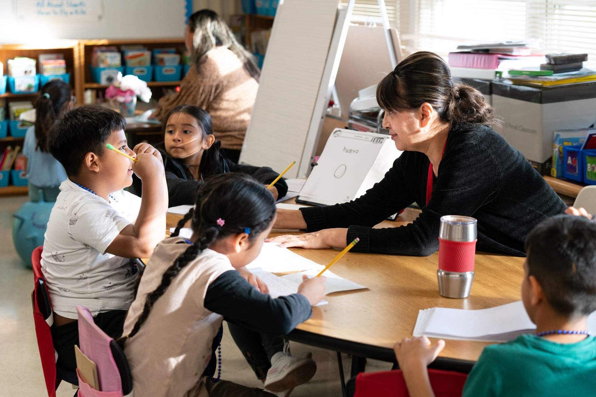A teacher assists children seated around a table, providing guidance and materials for their tasks.