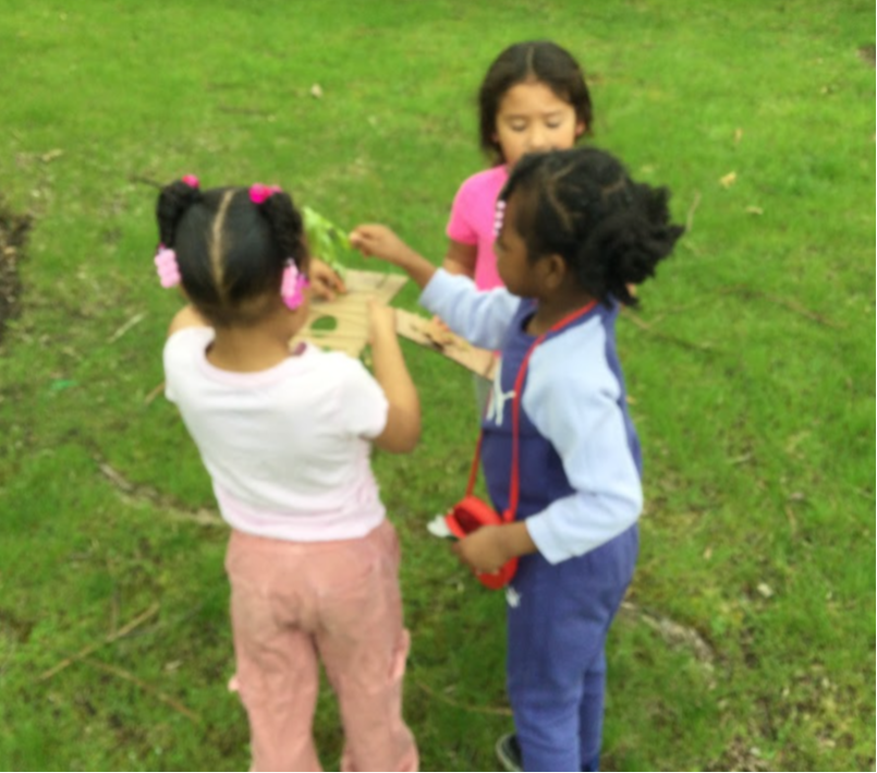 Three young children outside on a lawn. One child holds a piece of paper and another child holds a small green plant.