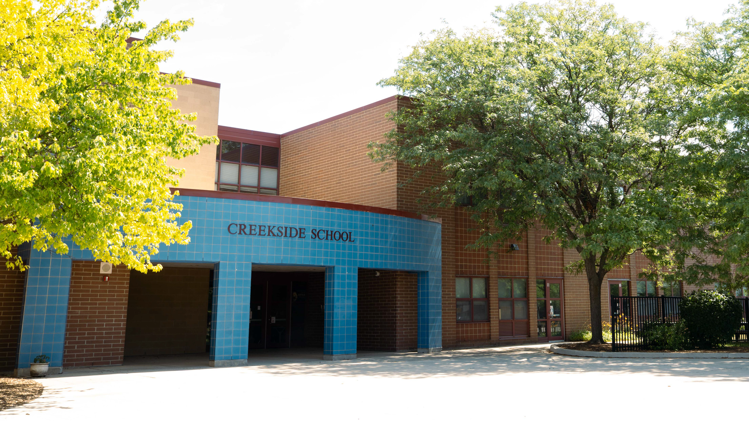 Blue and brown building with "Creekside School" text above two entrances. Trees, plants, and a fence behind.