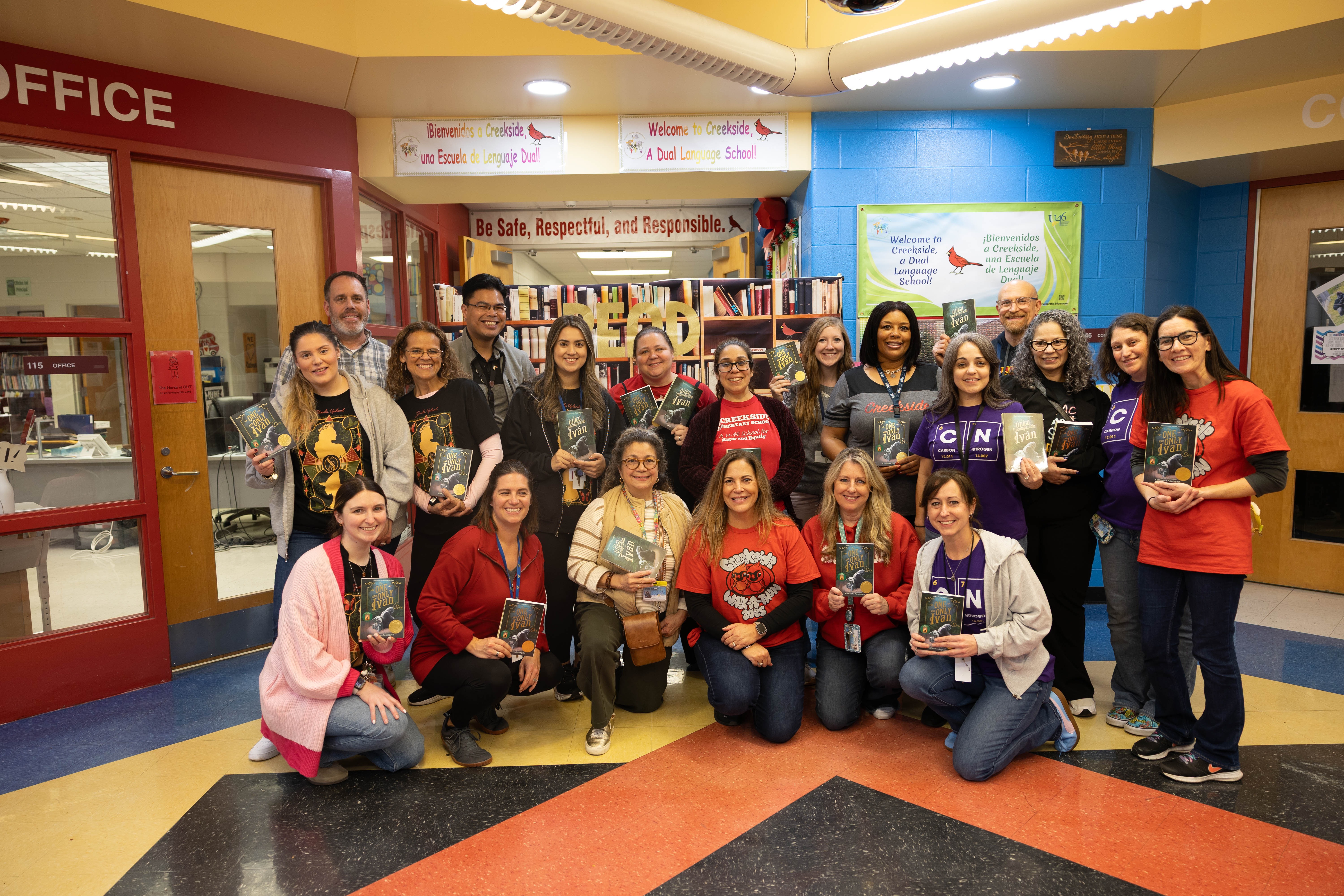 Group of people in a school hallway, holding books, smiling. Two doors in the background, shelves with books, and decorative walls.