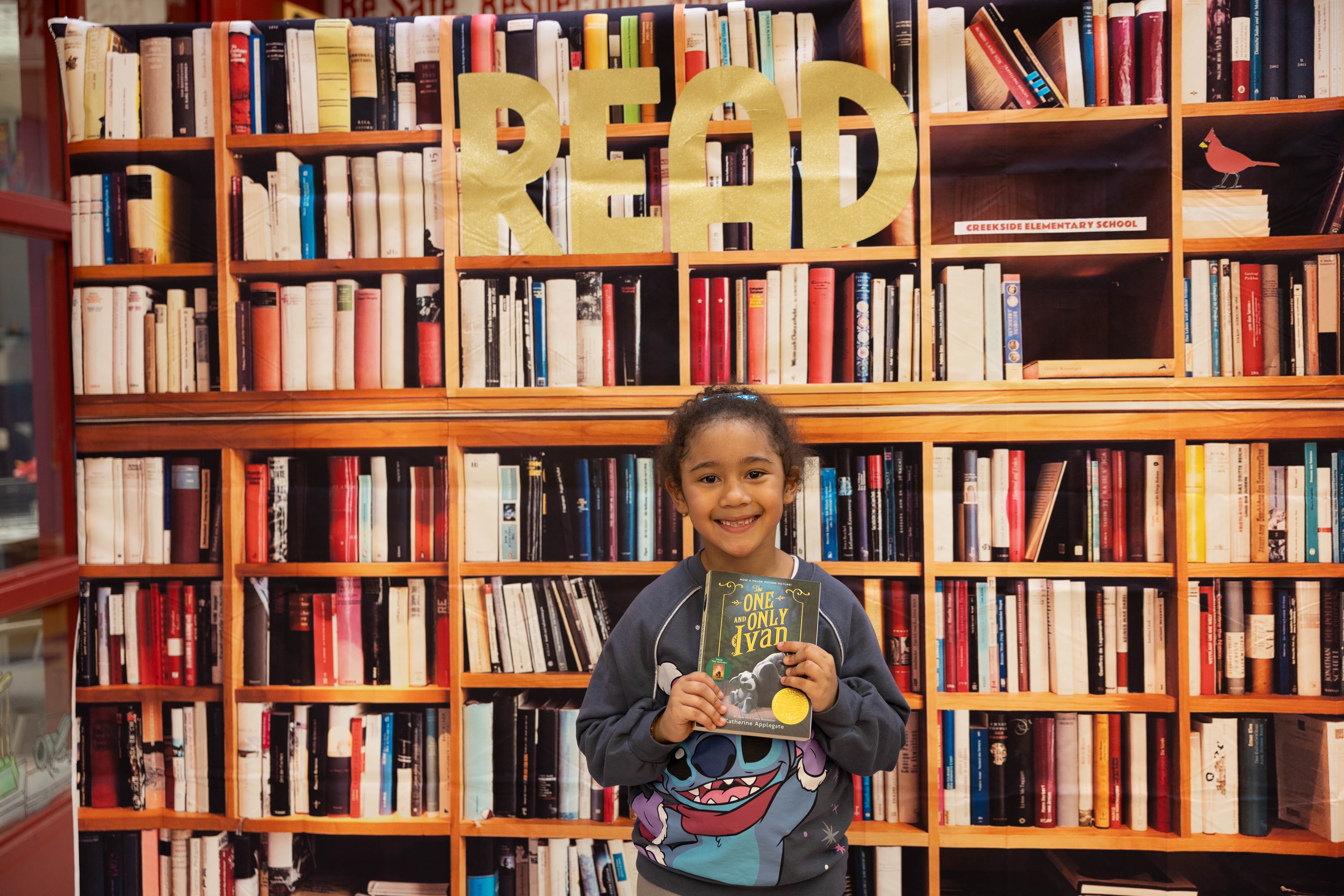 Child holds book in front of wall with large "READ" letters. Behind child is a tall bookshelf filled with many books.