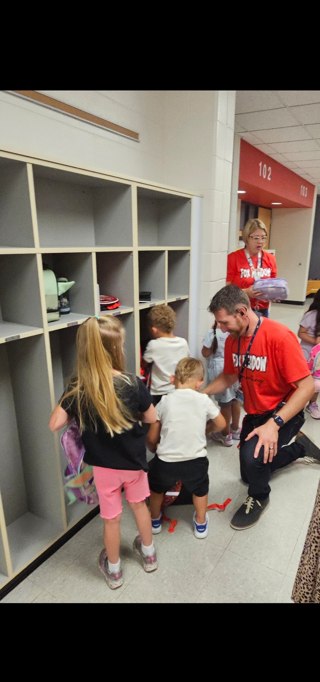 Adults and children interact by a row of lockers. A man kneels, holding a young boy. Nearby, a woman holds a bag.