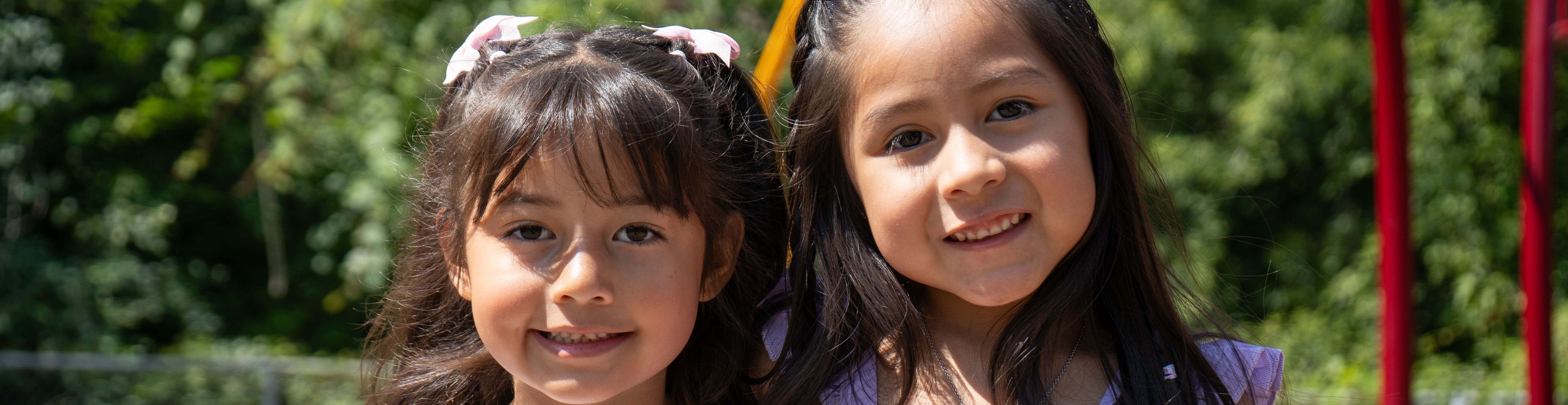 Two young girls stand in an outdoor playground area. One wears a white shirt, and the other wears a purple dress.