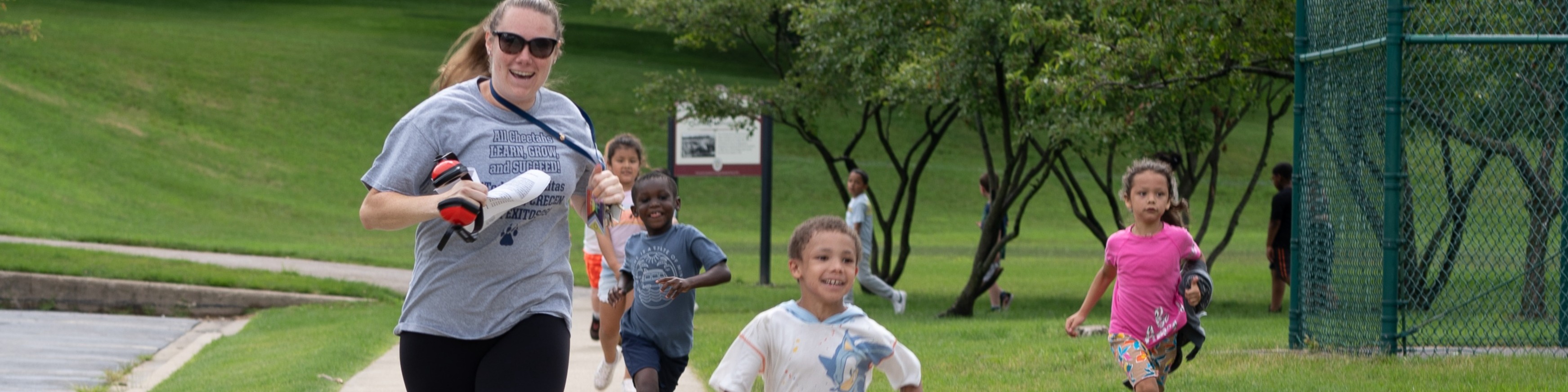 A woman and several children run along a sidewalk in a park with green grass and trees.