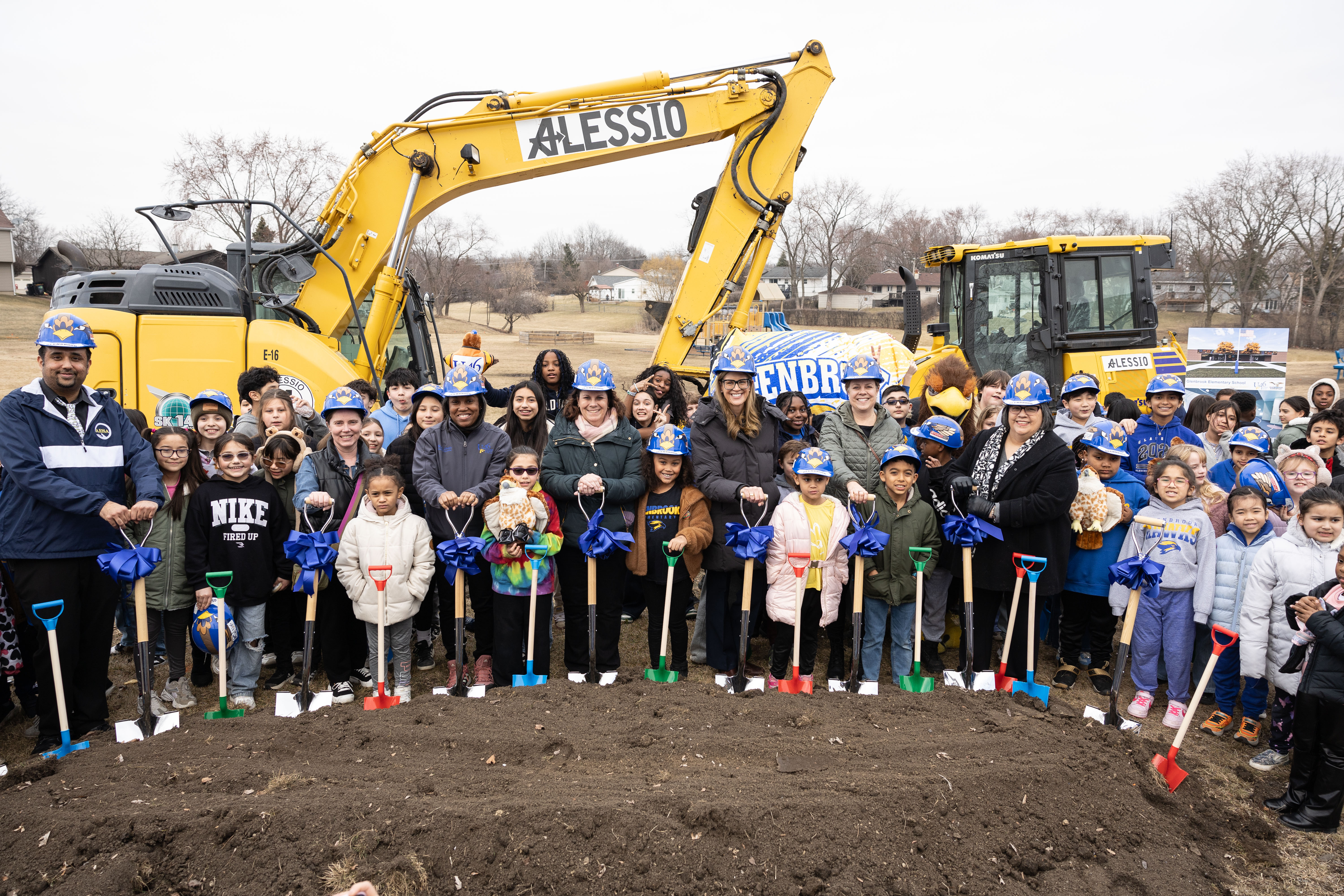 A group of people, including adults and children, stand in a circle holding shovels. Two yellow excavators are in the background.