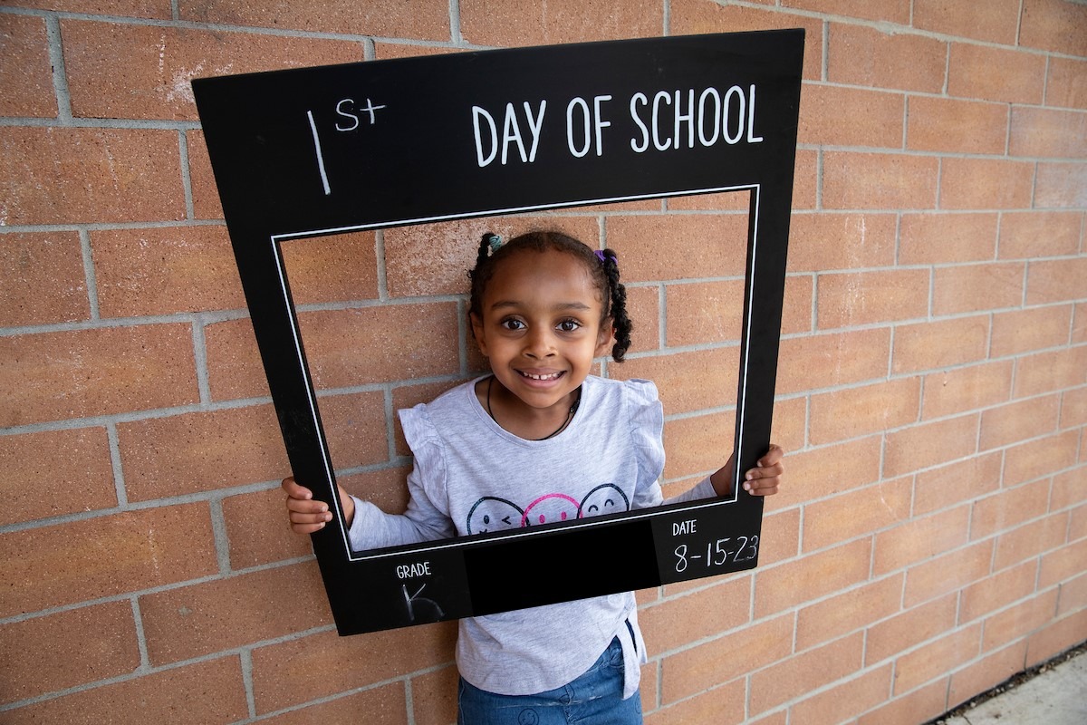 A young girl in a white shirt stands by a brick wall, holding a sign that says "Day of School".