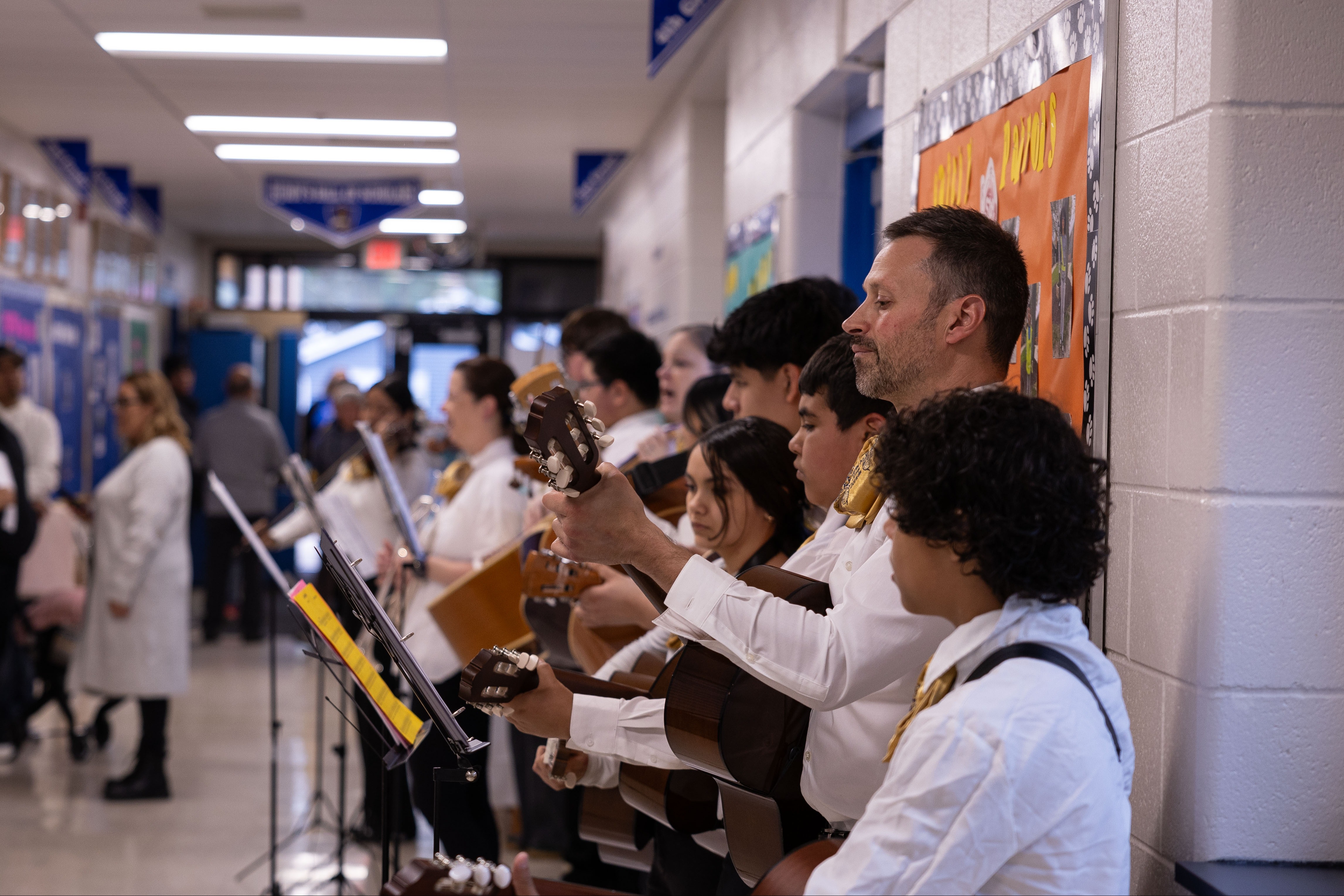 A group of musicians in white shirts play guitars in a hallway; some people stand in the background.
