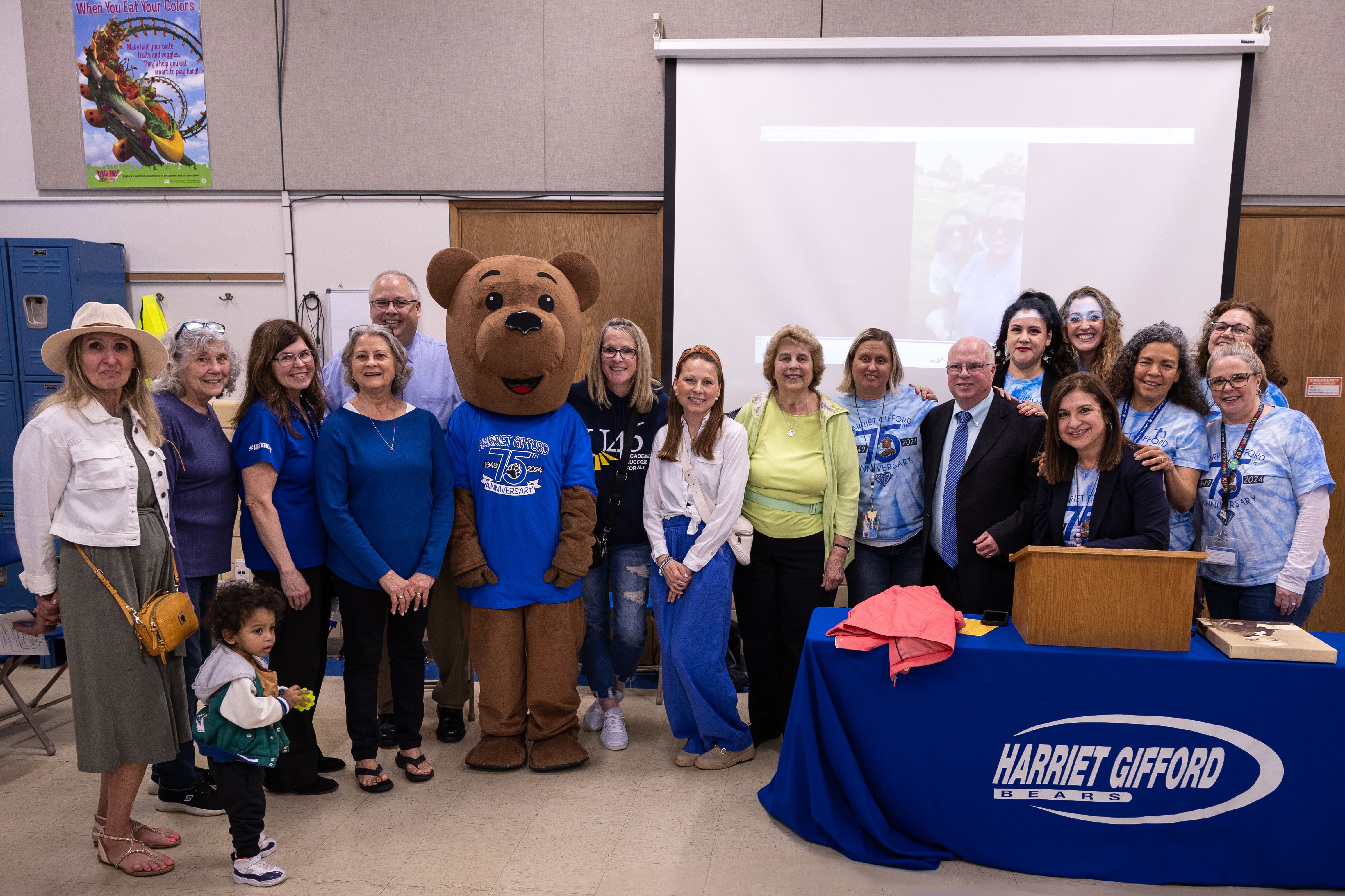 A group of people in blue shirts stands with a mascot in a room. A podium with a logo is nearby.