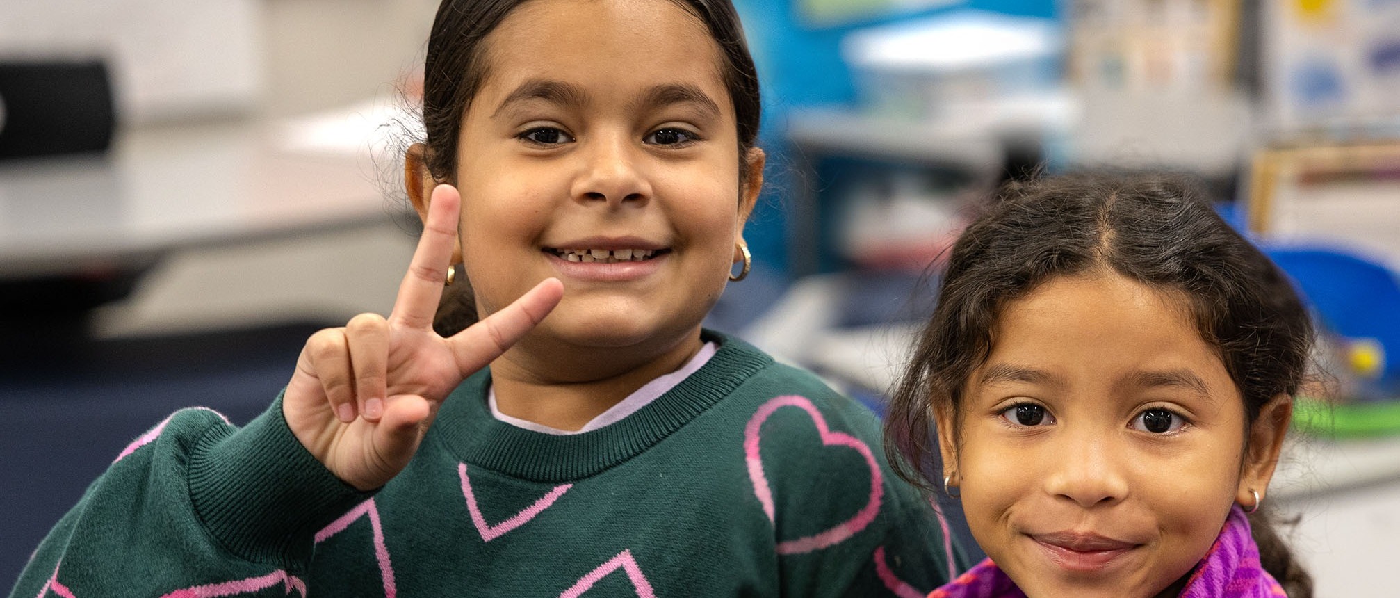 Two girls in a classroom, one wearing green with pink hearts and the other in purple. They smile and pose.