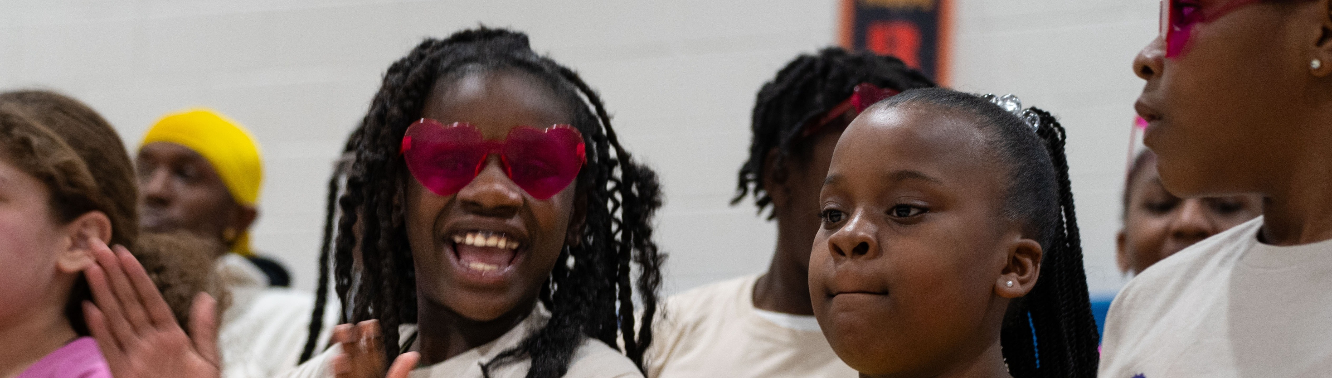 Several children clap hands in a room. They wear white shirts, and one girl wears pink.