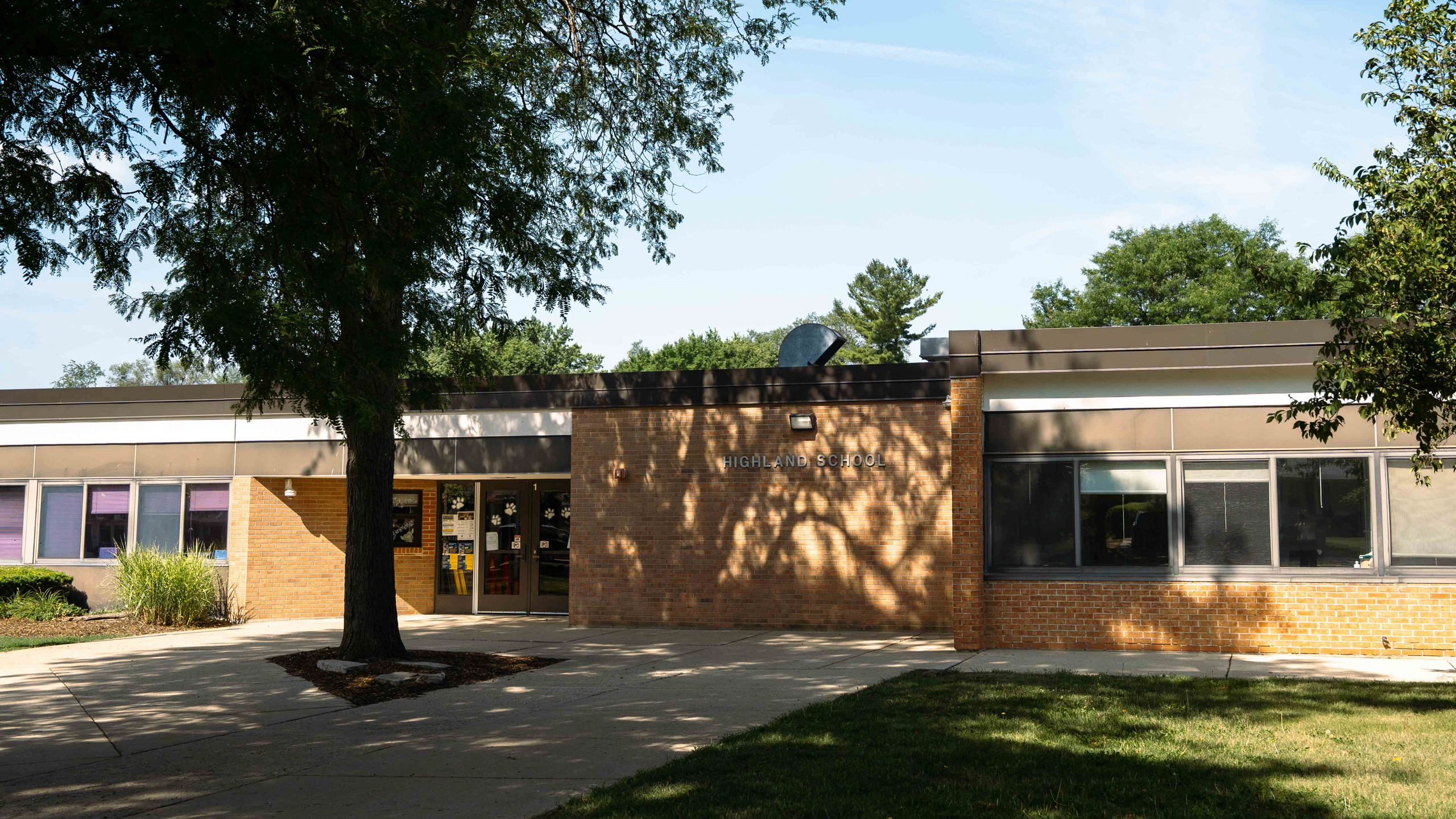 Exterior of a school building. Brick walls, glass windows, and a tree in front. Shaded walkway leads to entrance.