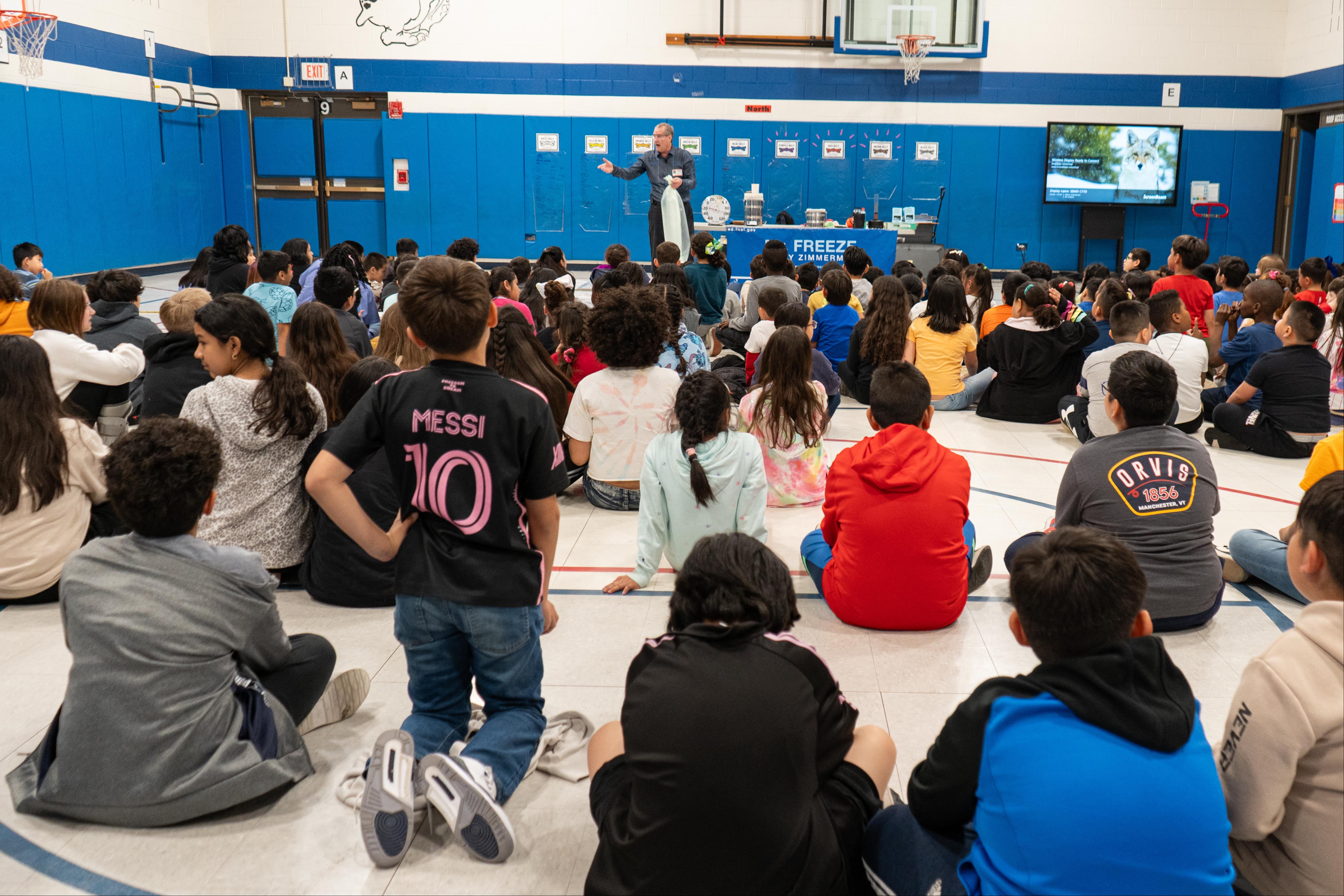 Several students gather in a gym. A man in a black shirt stands before them.