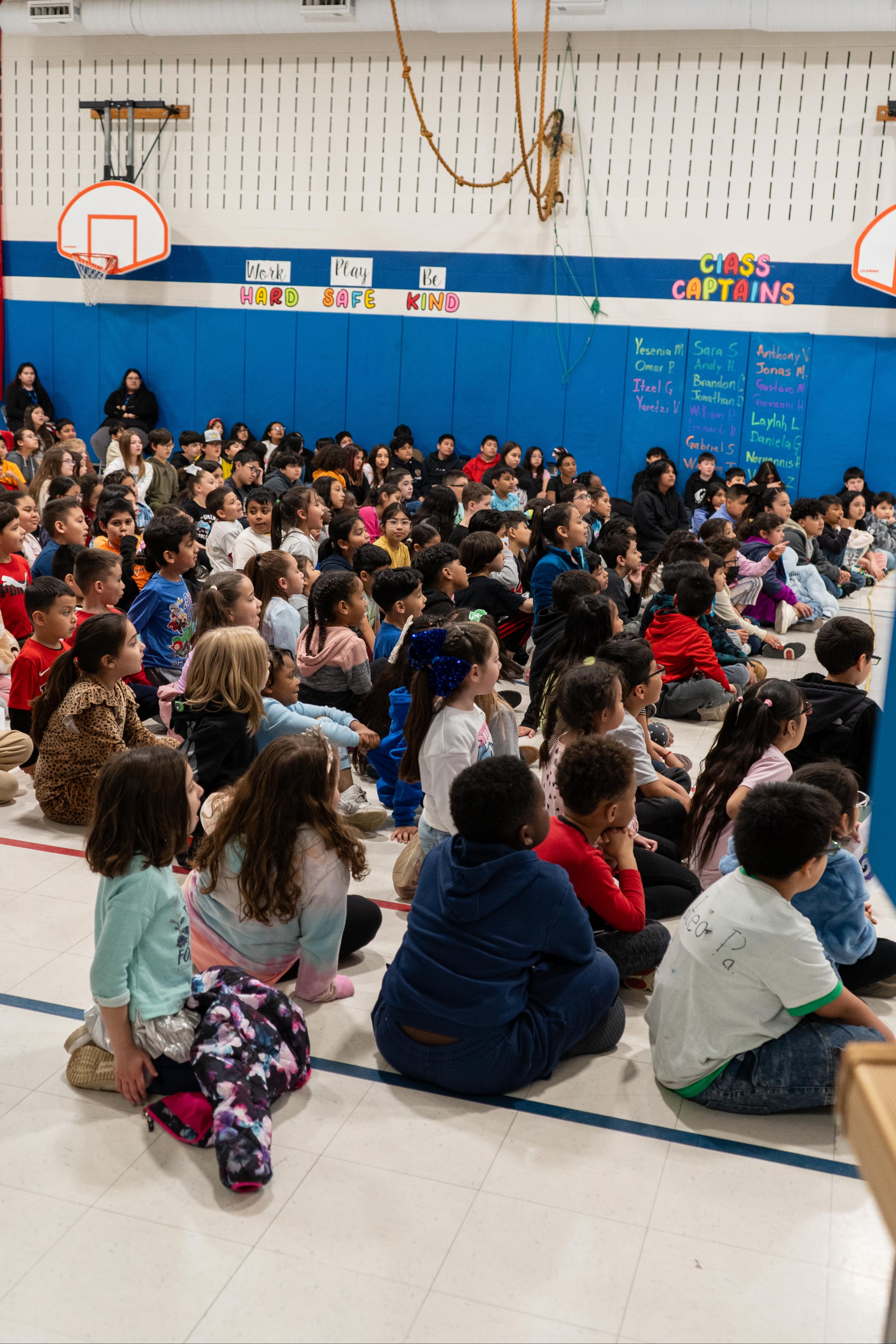 Indoors, many children sit on a floor in front of a wall with a basketball hoop.