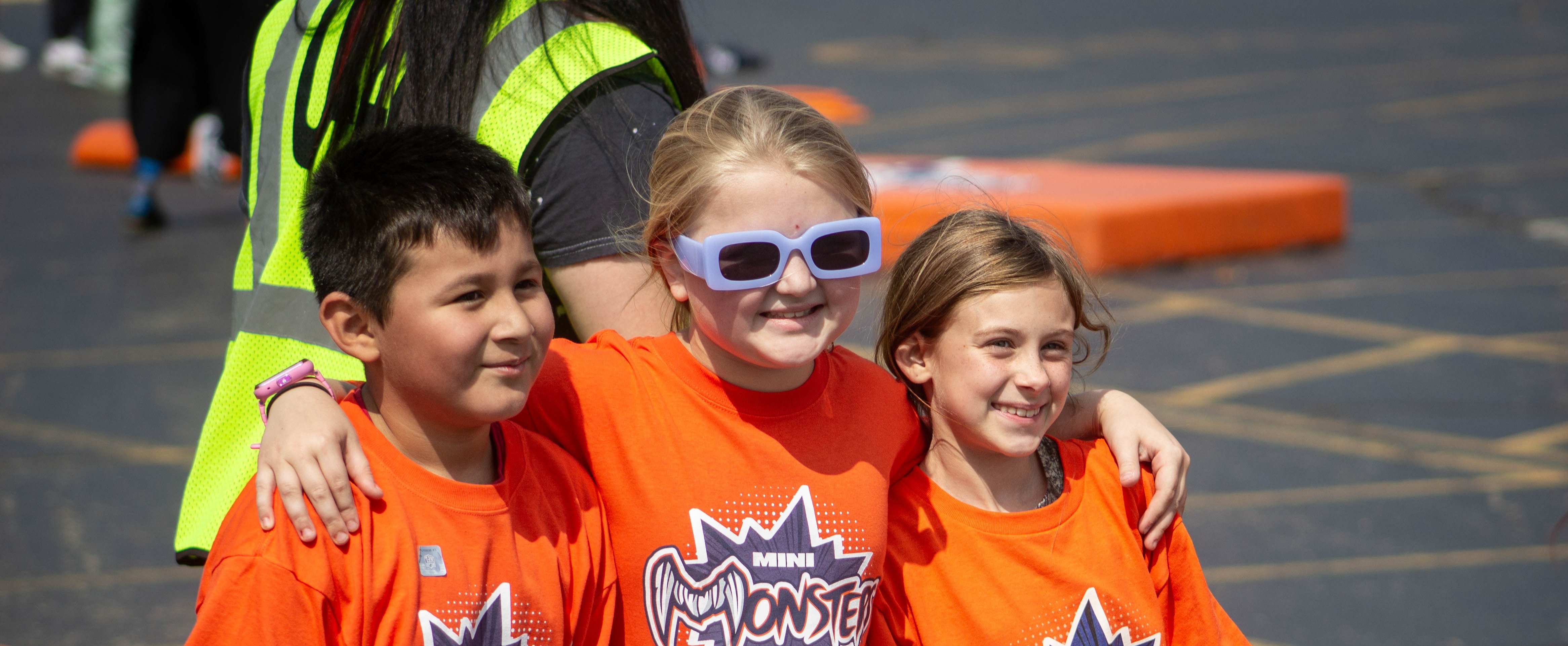 Three children in orange shirts stand together. One wears sunglasses and a smile, while two wear yellow lanyards.